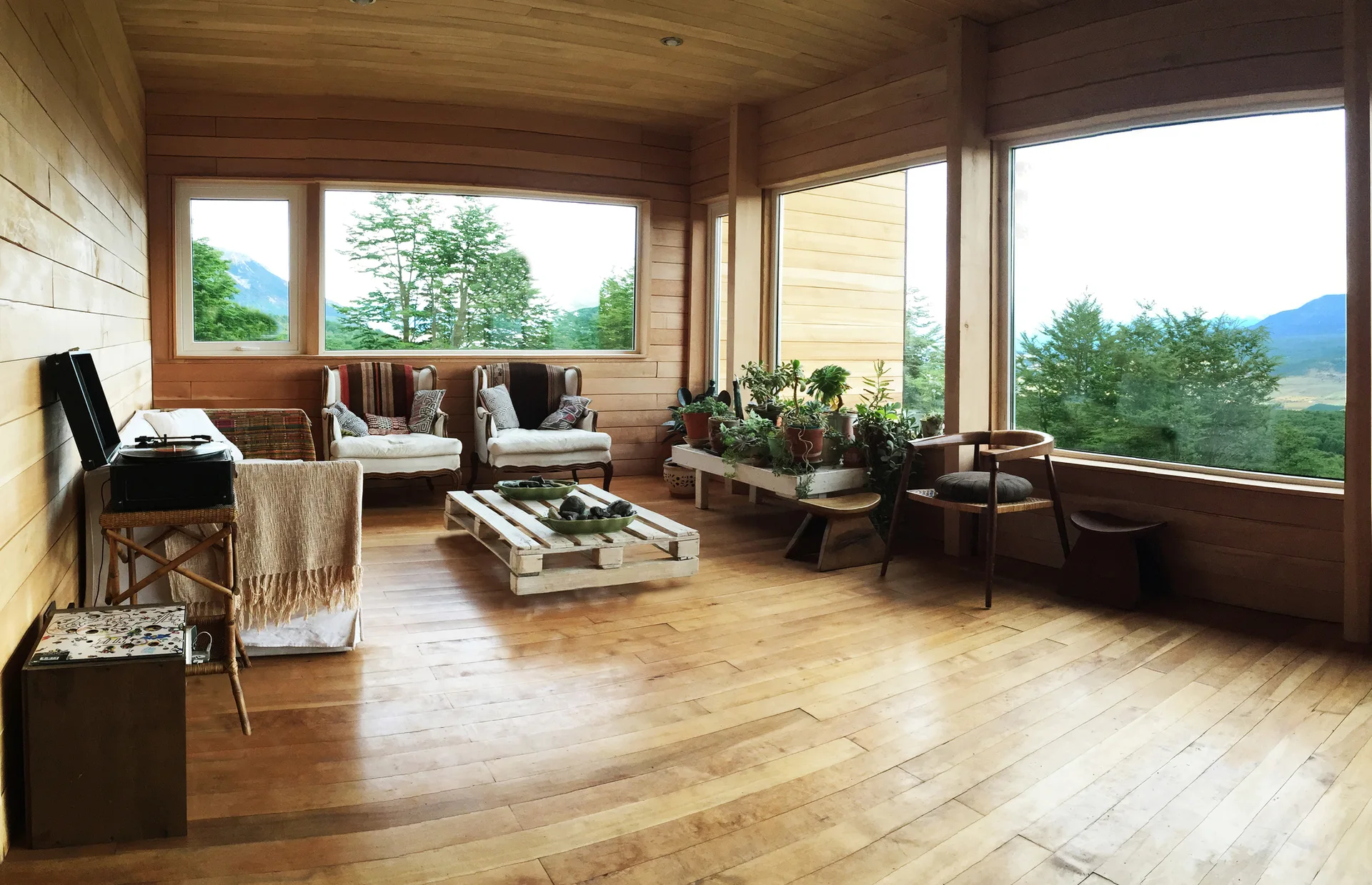 A rustic living room wrapped in warm wood paneling features a low pallet coffee table, neutral armchairs, and large windows looking out on green trees.