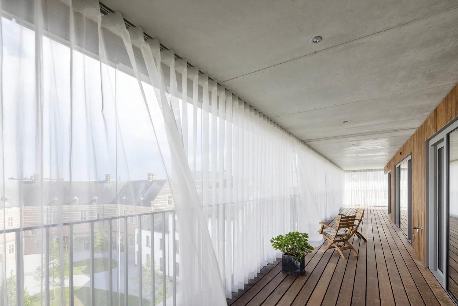 A long outdoor balcony features natural wood plank flooring, a matching wood slat wall, concrete ceiling, and long sheer white curtains.