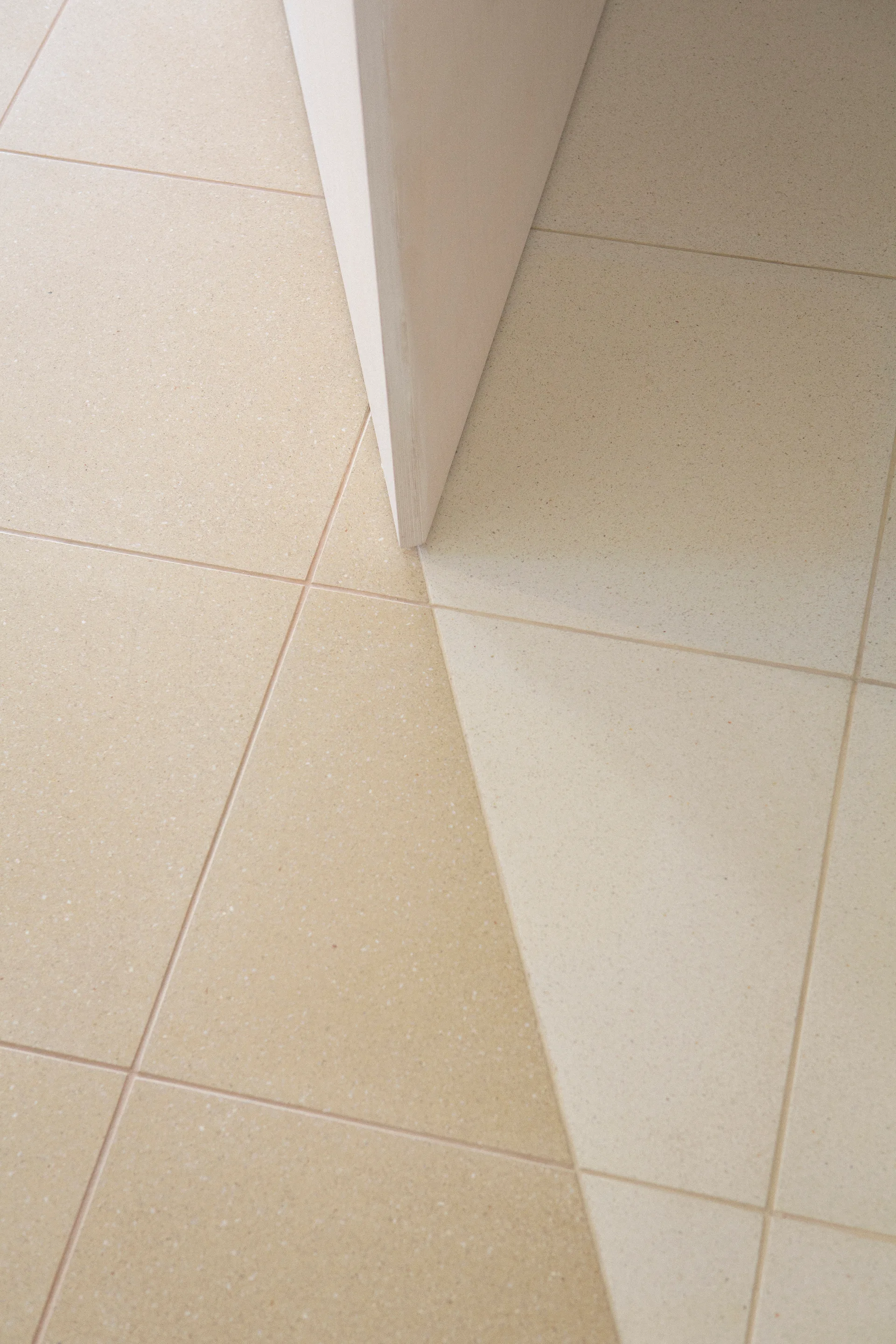 Close-up of matte speckled floor tiles featuring a diagonal color change from beige to cream next to an off-white wall partition.
