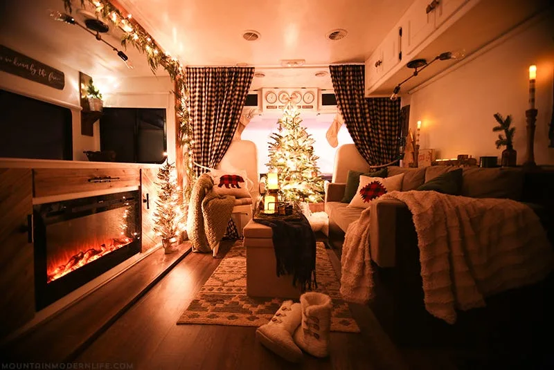 An RV living room decorated for Christmas with an electric fireplace, a lit tree, buffalo check curtains, and warm ambient lighting.