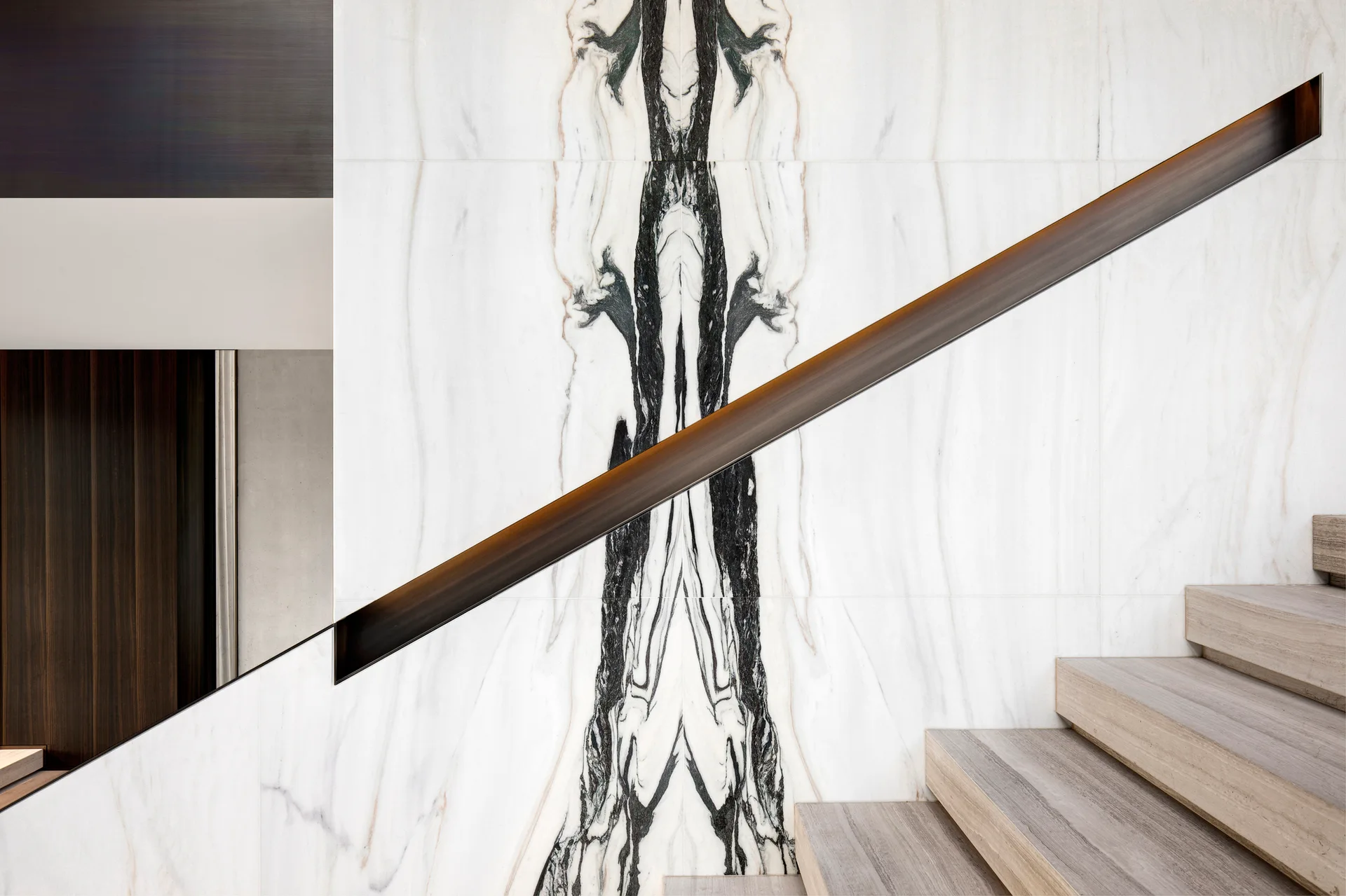 A modern staircase with light wood treads climbs alongside a bookmatched marble wall featuring a recessed diagonal metal handrail with subtle lighting.
