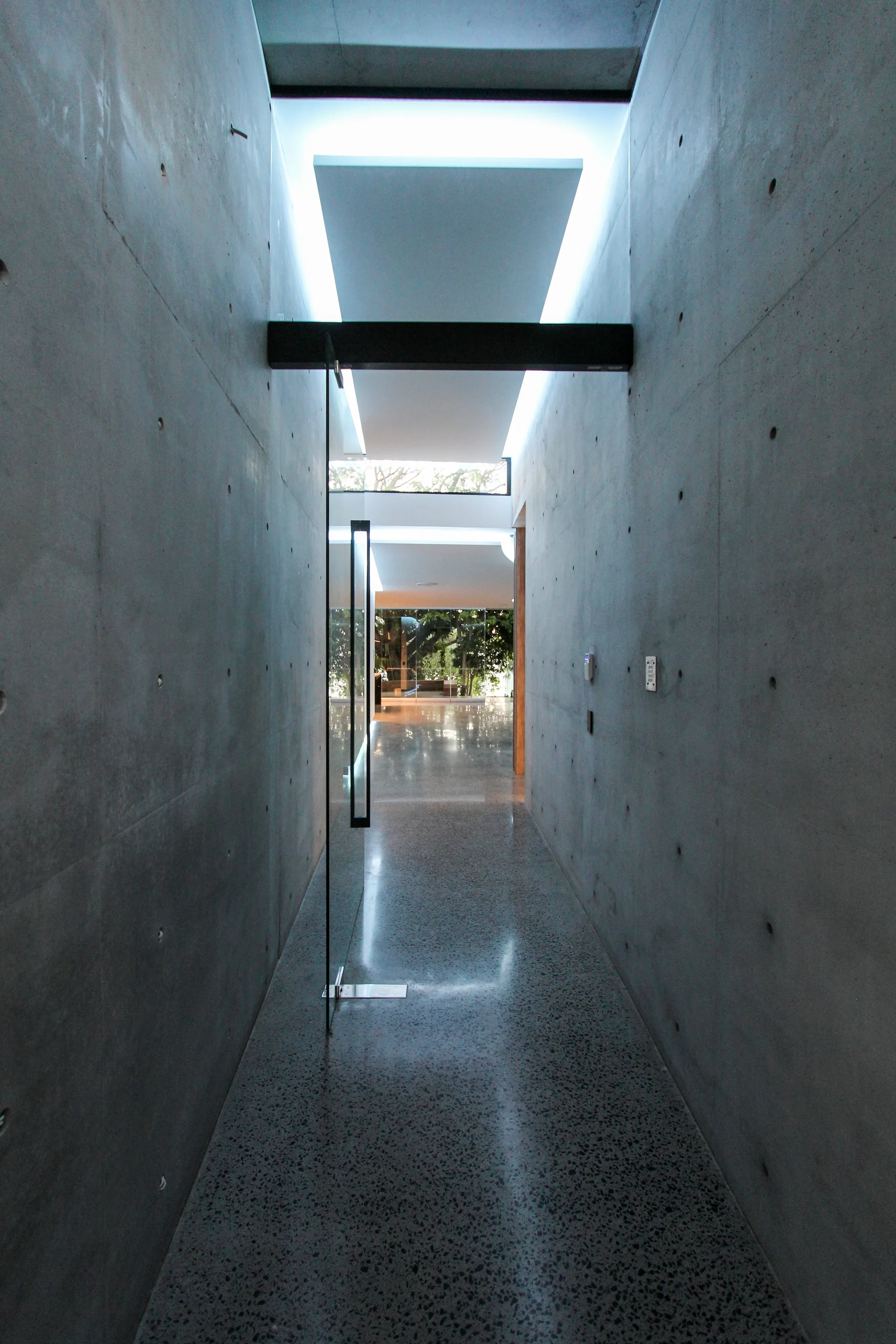 A long, modern hallway featuring exposed concrete walls with circular tie holes, a polished speckled terrazzo floor, and a tall glass door.
