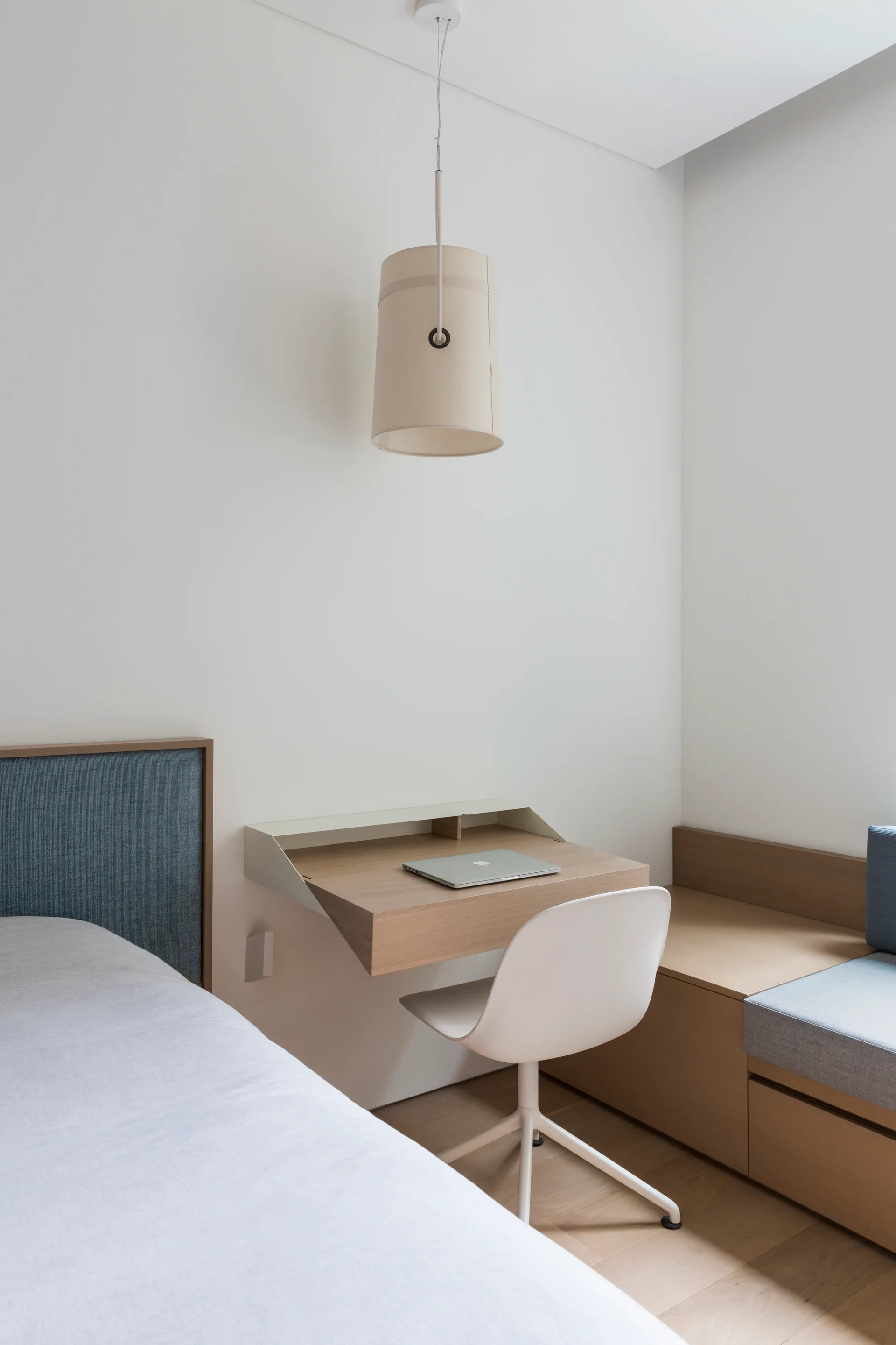 A minimalist bedroom features a floating wooden desk, a white swivel chair, a fabric pendant light, and an integrated bench beside a bed.