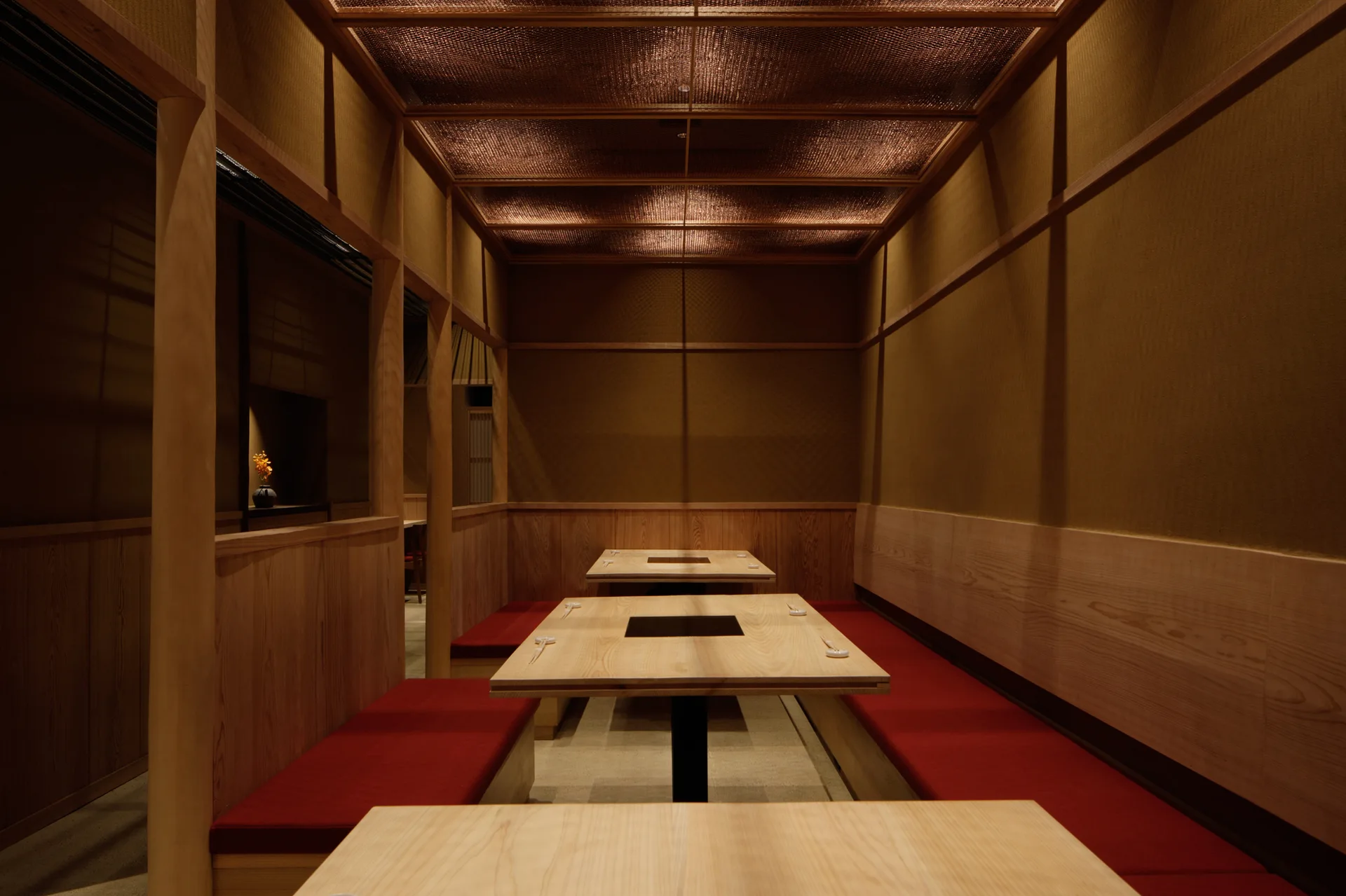 Long light-wood dining tables sit between built-in wooden benches with red cushions beneath a backlit grid ceiling.