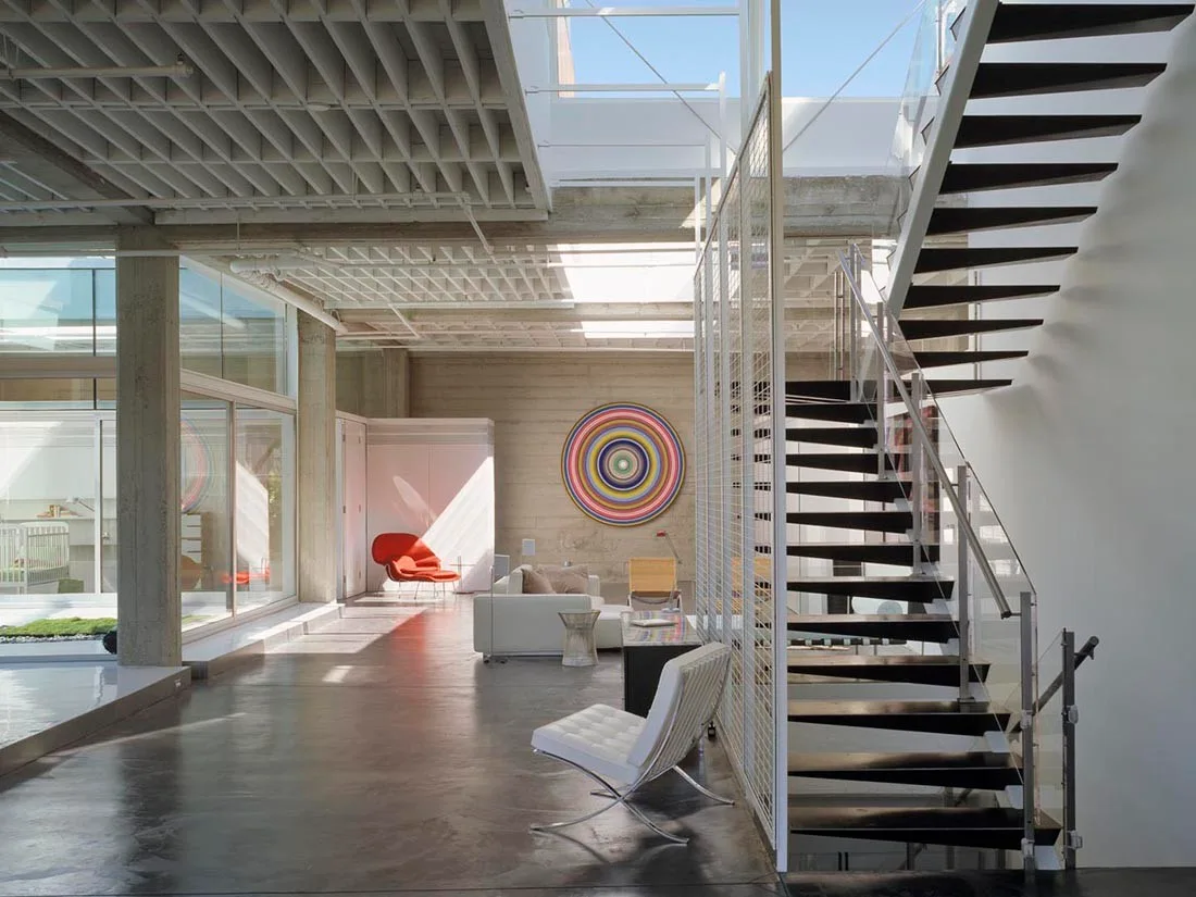 A spacious industrial living room features a dark floating staircase, concrete waffle ceiling, white sectional sofa, and polished dark concrete floors.
