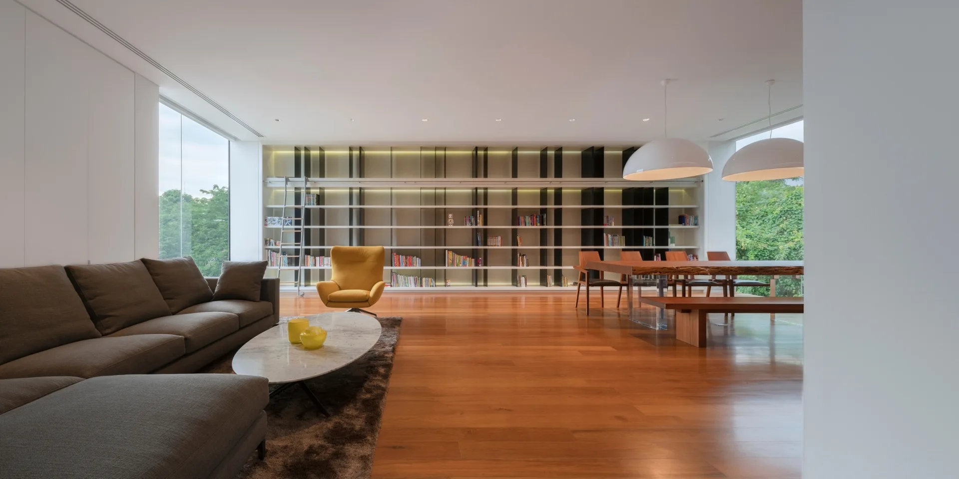 A spacious living room features a floor-to-ceiling library wall with a rolling ladder, a grey sectional sofa, and a wood dining table.