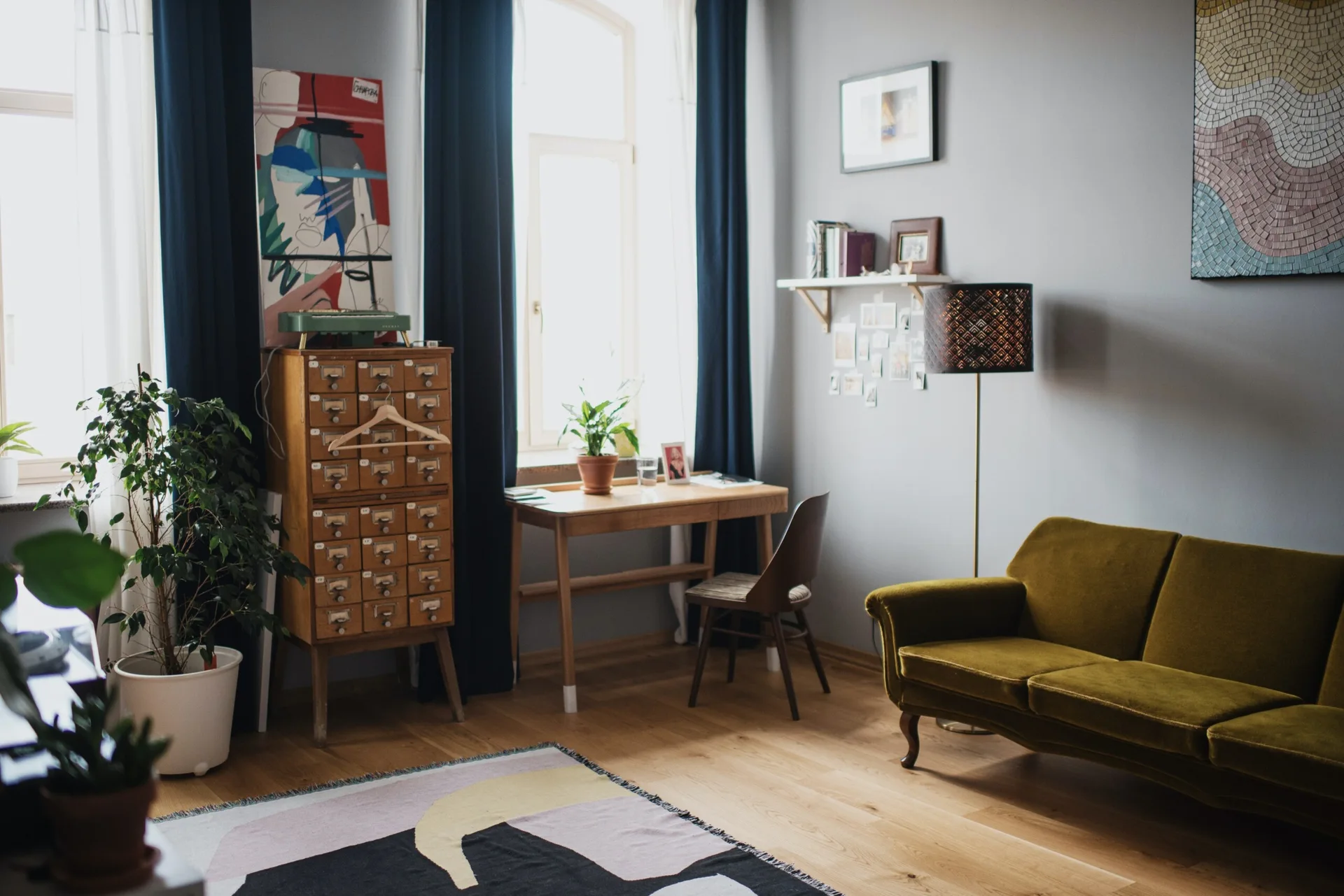 A living room featuring an olive green velvet sofa, a vintage wooden library cabinet, and a writing desk positioned in front of a window.