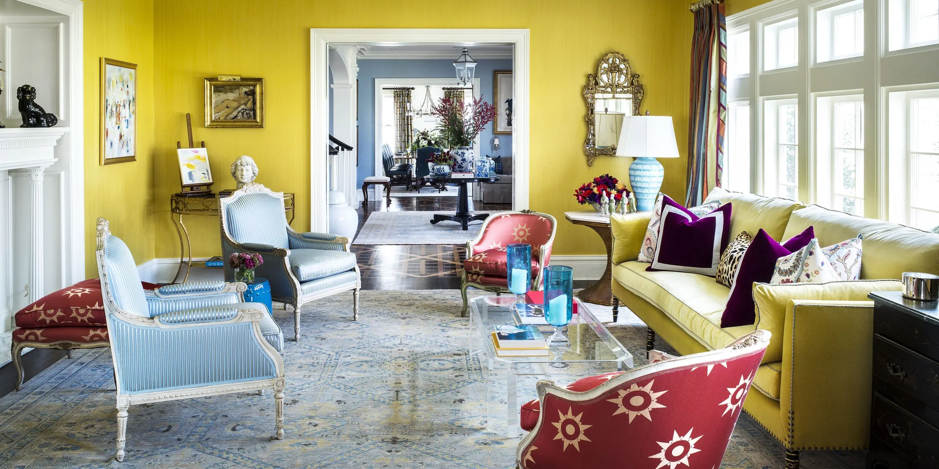A yellow living room features a yellow sofa, blue striped armchairs, red patterned armchairs, and a clear acrylic coffee table on a large rug.