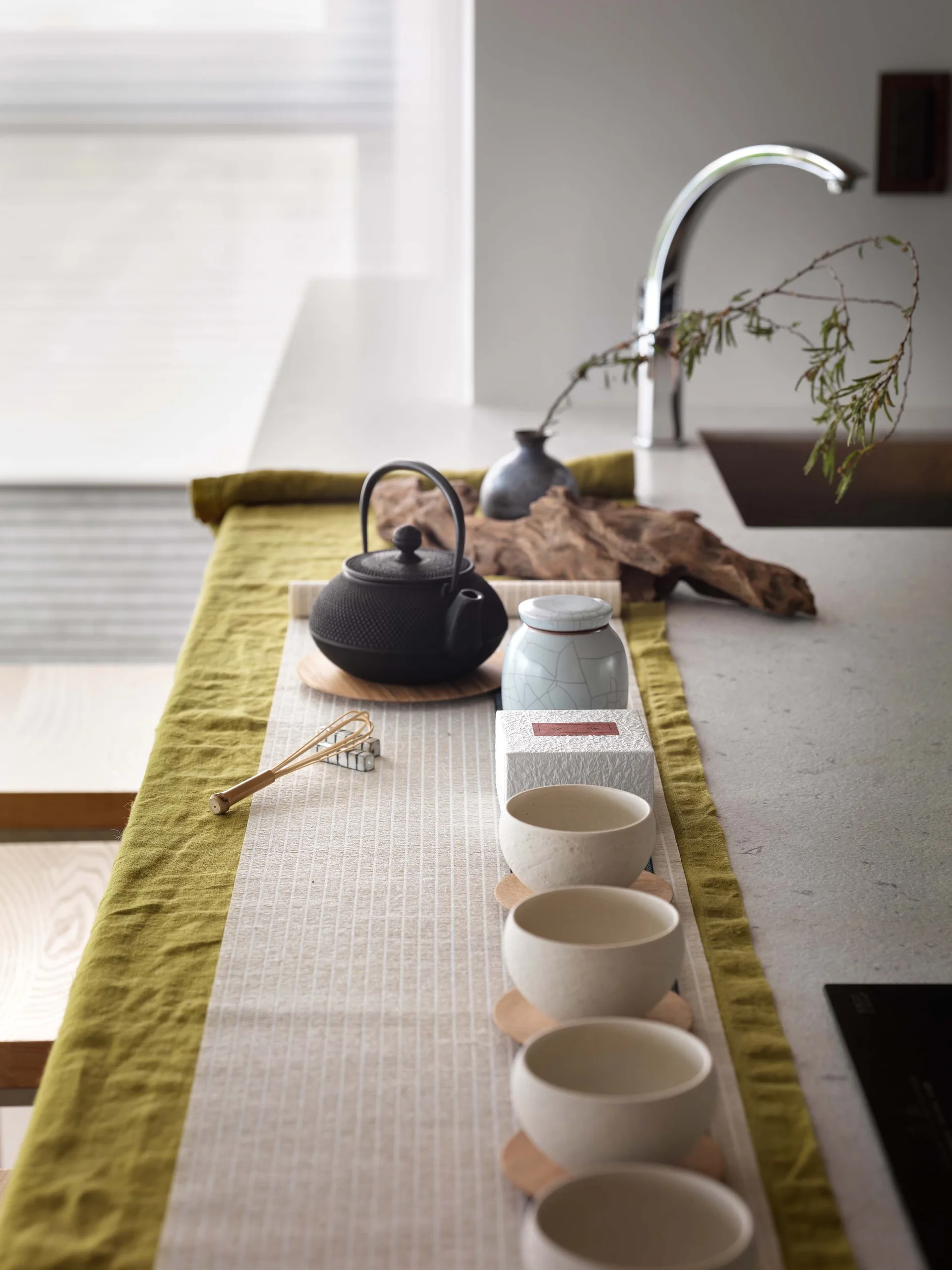 A cast iron teapot, ceramic cups, and a matcha whisk are arranged on an olive green fabric runner on a stone kitchen countertop.