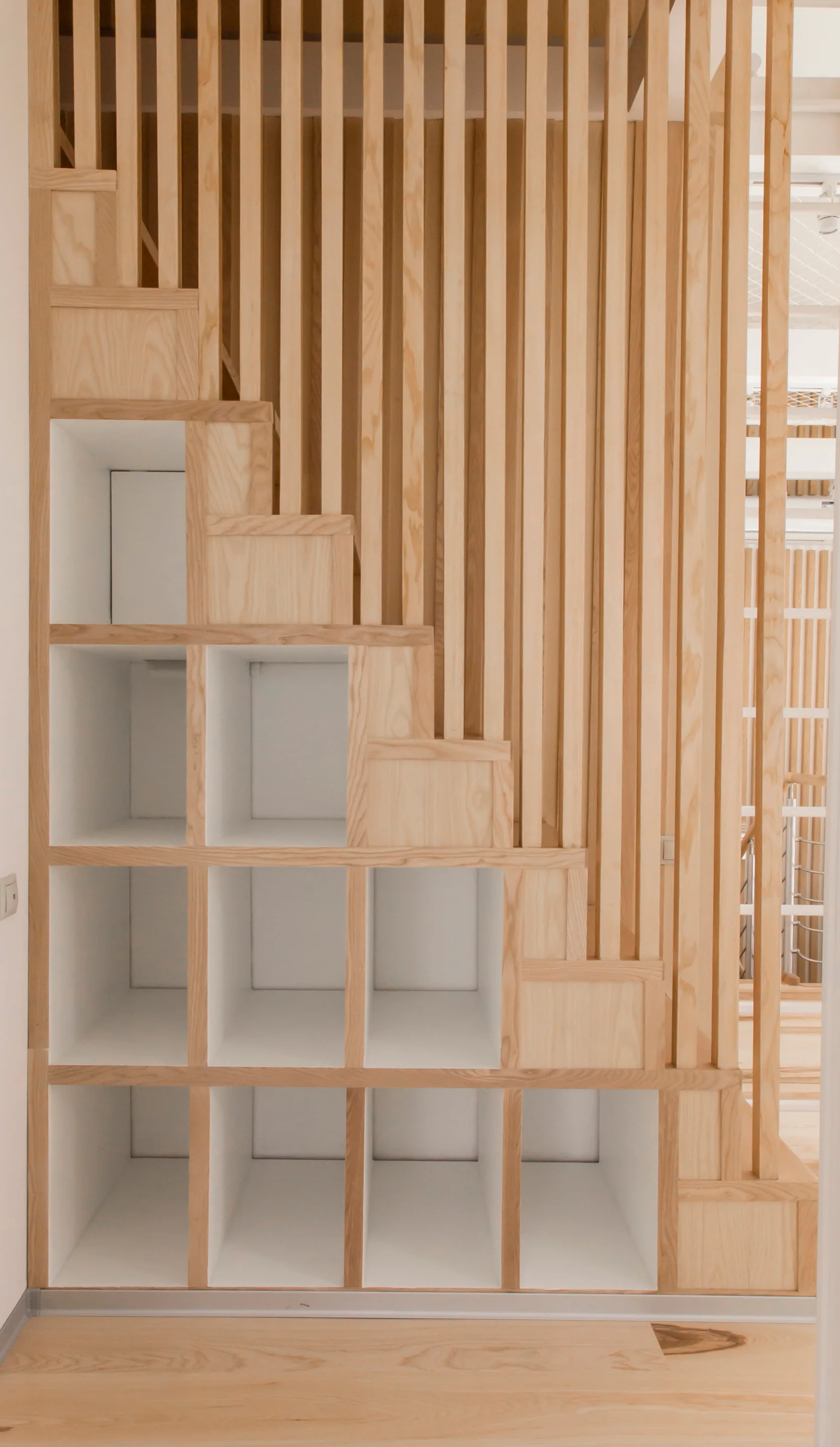 A light wood staircase featuring integrated square white open shelving compartments underneath and a tall partition screen of vertical wooden slats.