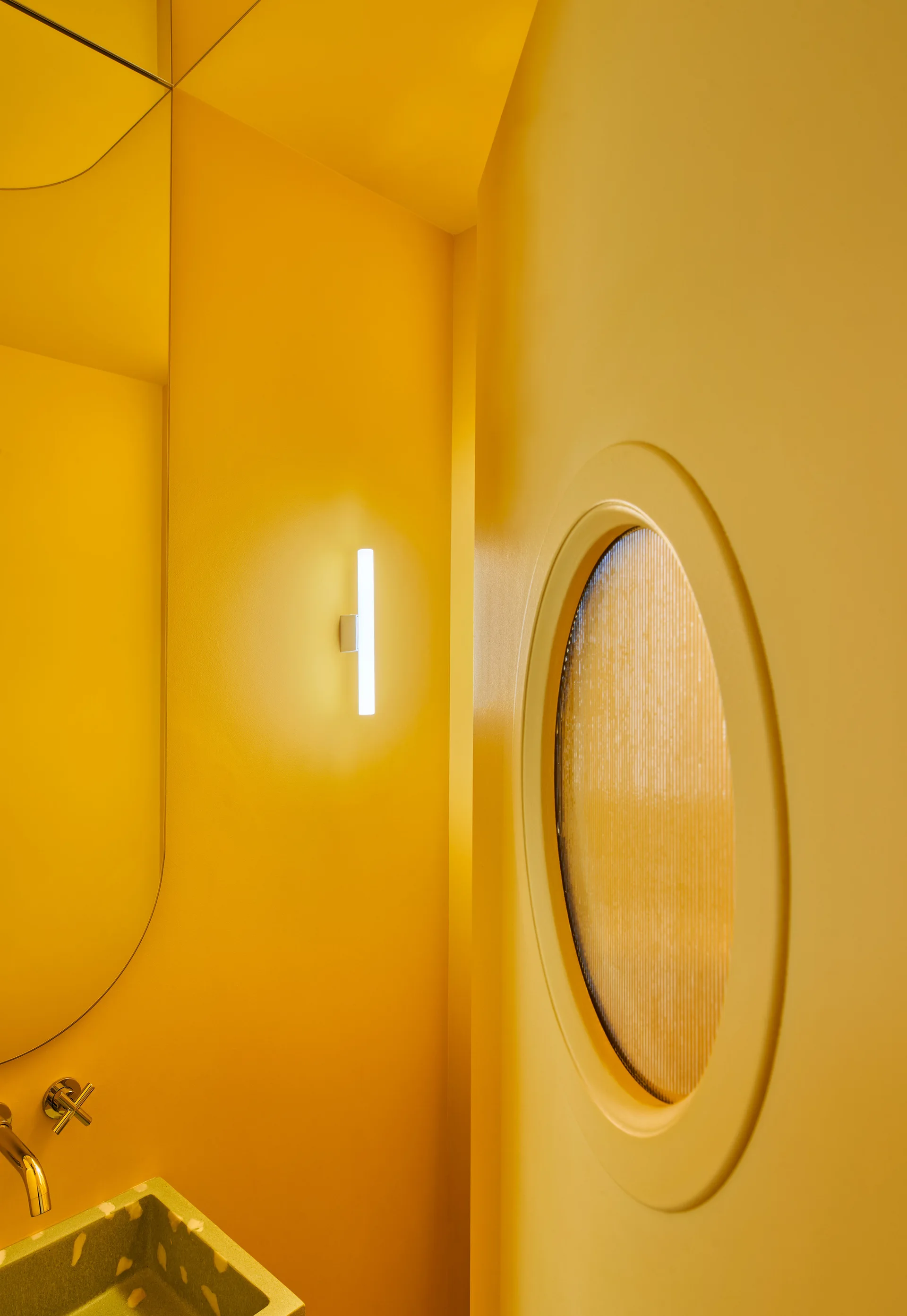 A bright yellow powder room corner features a round porthole window with ribbed glass, a glowing linear wall sconce, and a matching yellow wall.
