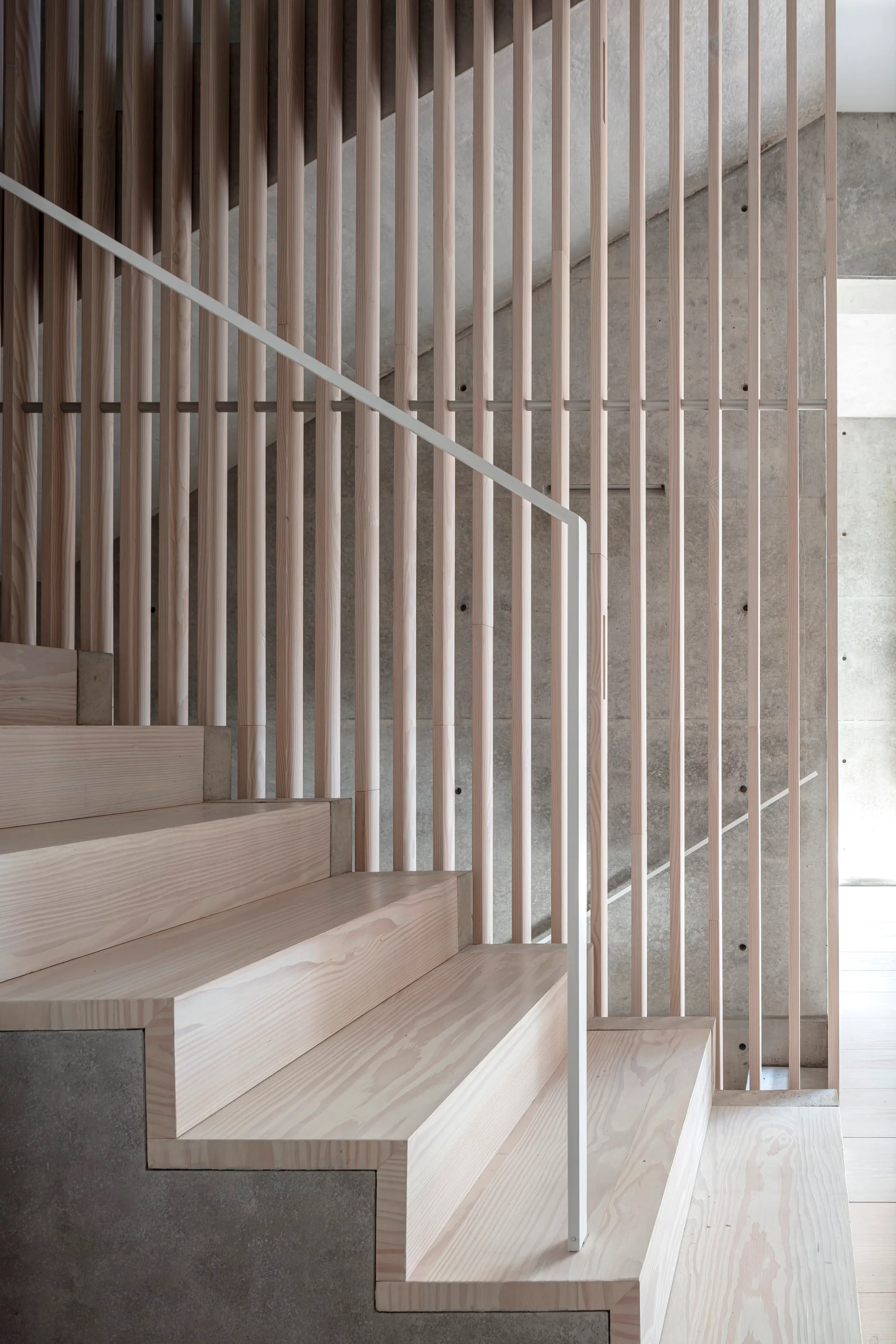 A minimalist staircase features light wood steps, a thin white handrail, an exposed concrete wall, and a tall screen of vertical wooden slats.