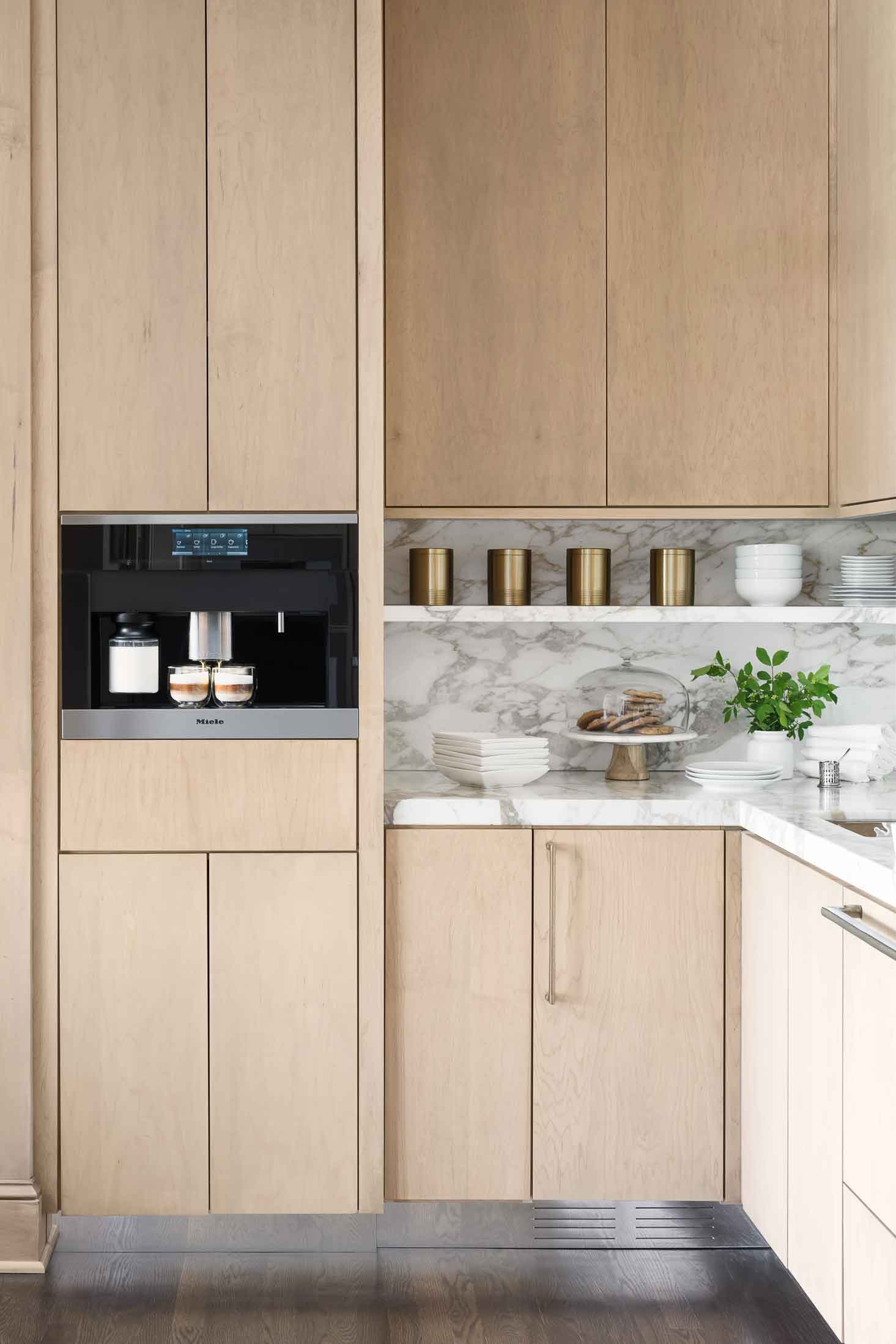 Modern kitchen features light wood slab cabinetry, a built-in espresso machine, a marble backsplash, and an open shelf displaying gold canisters.