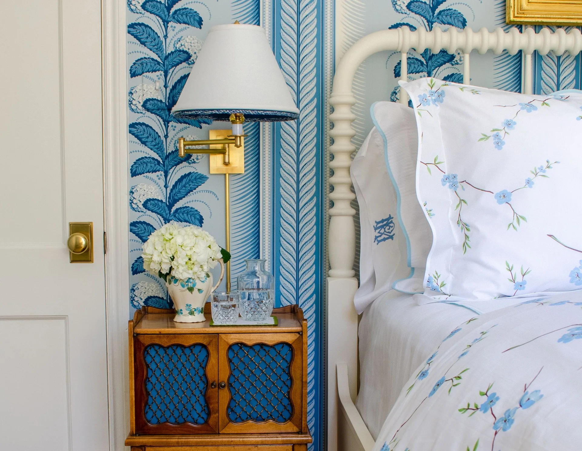 A traditional bedroom corner features a white spindle bed, a wood nightstand, and blue and white floral wallpaper with a brass sconce.