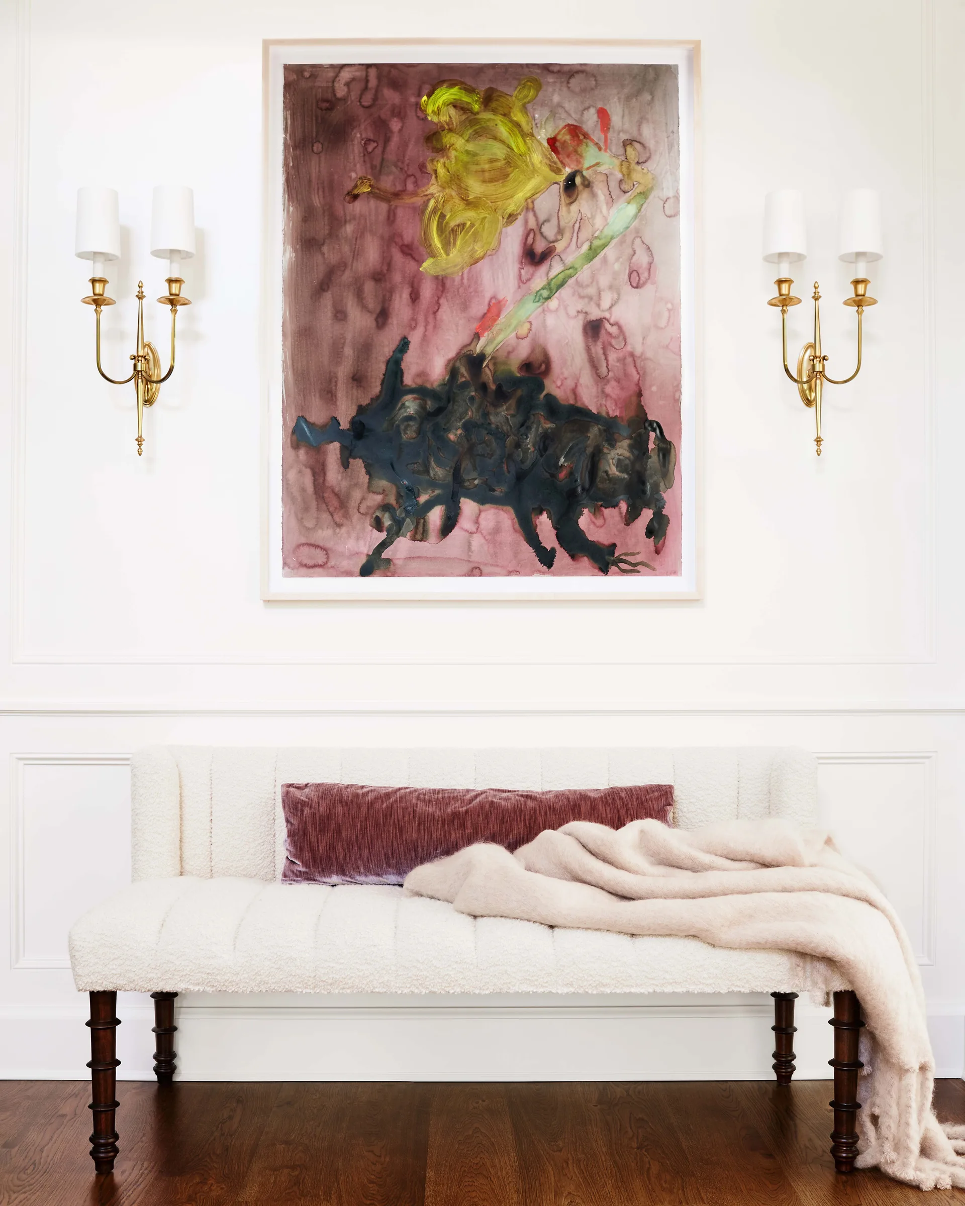 A cream textured bench sits below a large abstract painting flanked by symmetrical brass wall sconces on a white paneled wall.