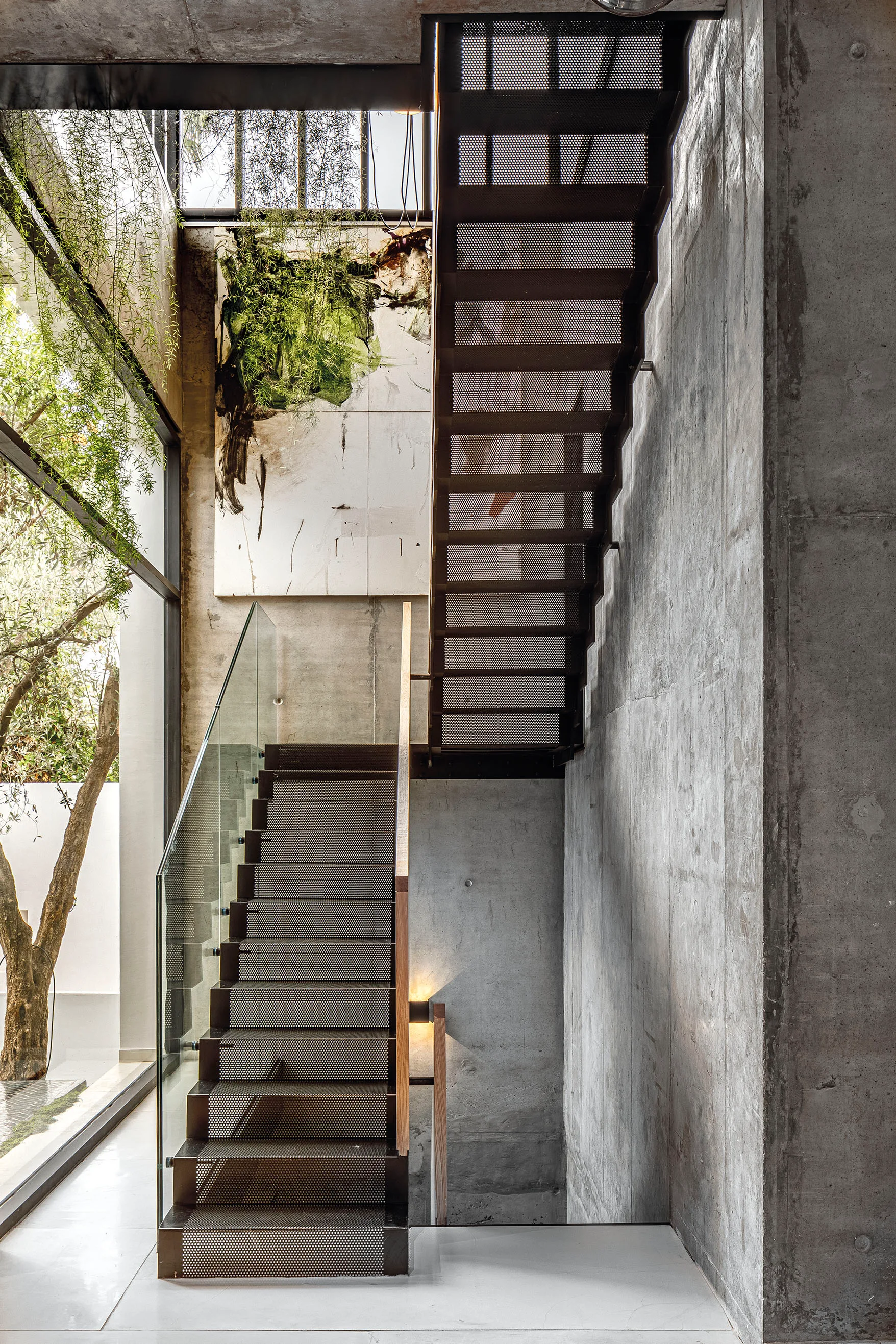 A modern industrial staircase with perforated steel steps and a glass railing is set against raw concrete walls next to a large window.