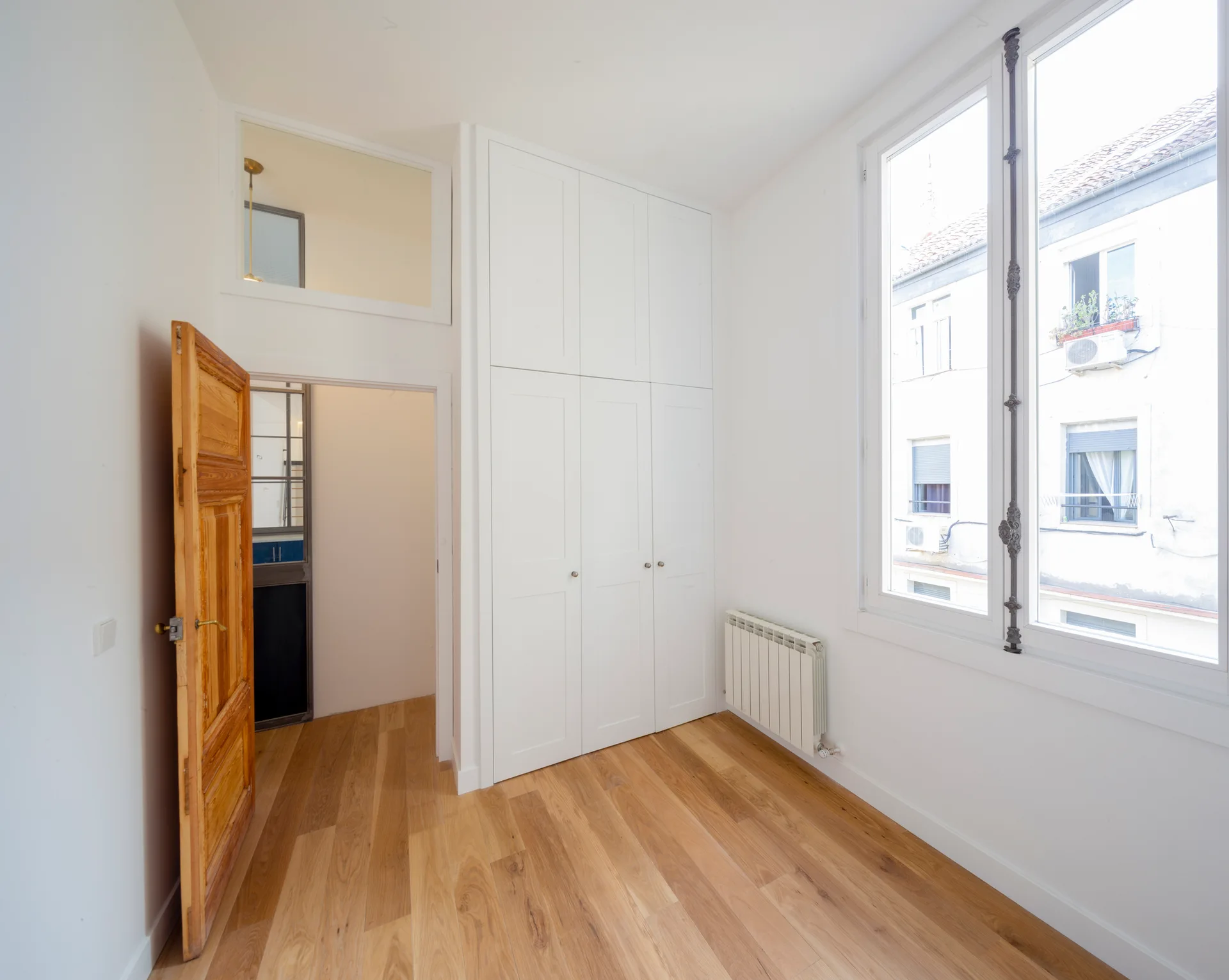 An empty bright room featuring light hardwood flooring, a tall open natural wood door, white built-in floor-to-ceiling closets, and a large window.