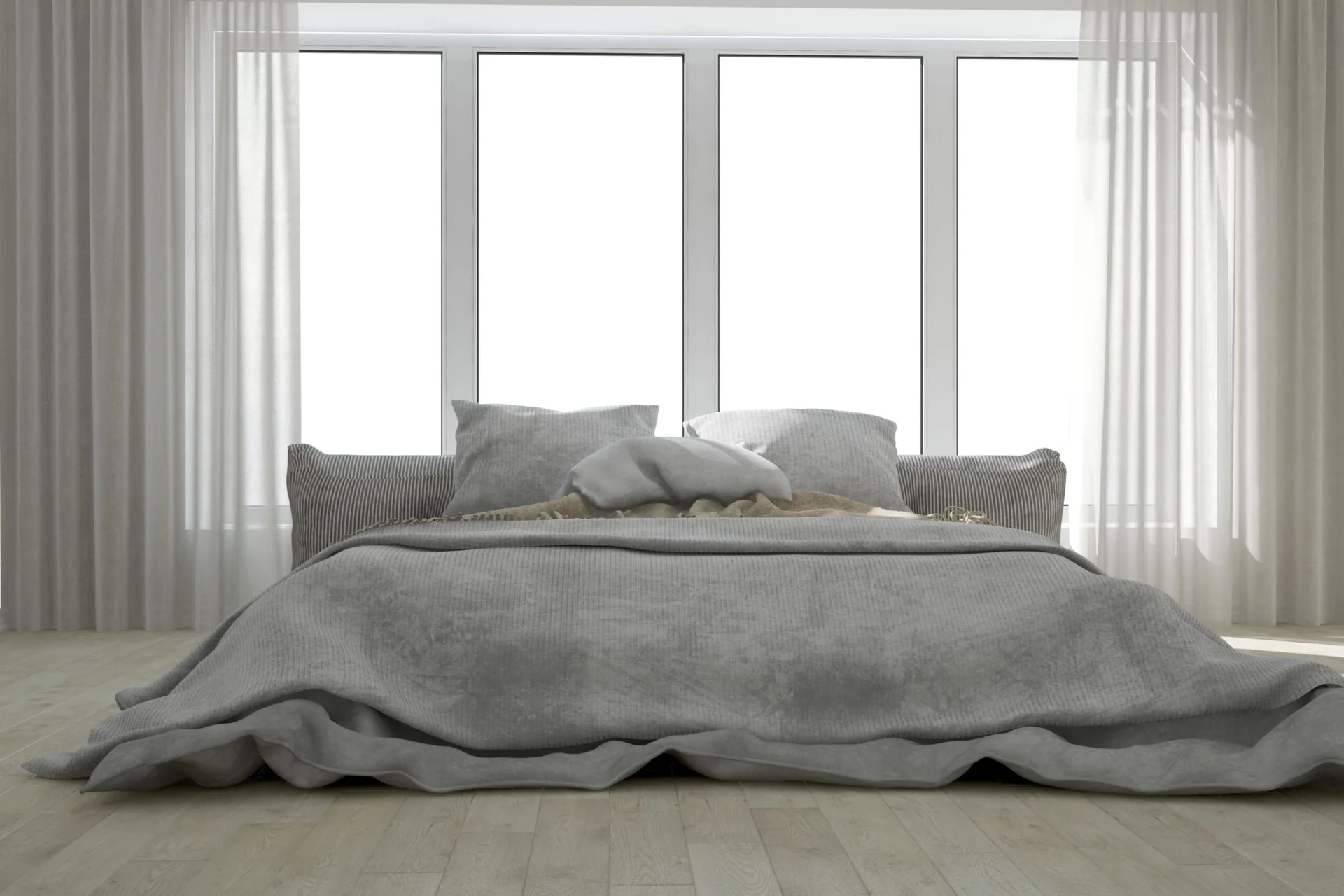 A low-profile grey bed with matching linens sits in front of a wide window framed by sheer white curtains on light wood floors.