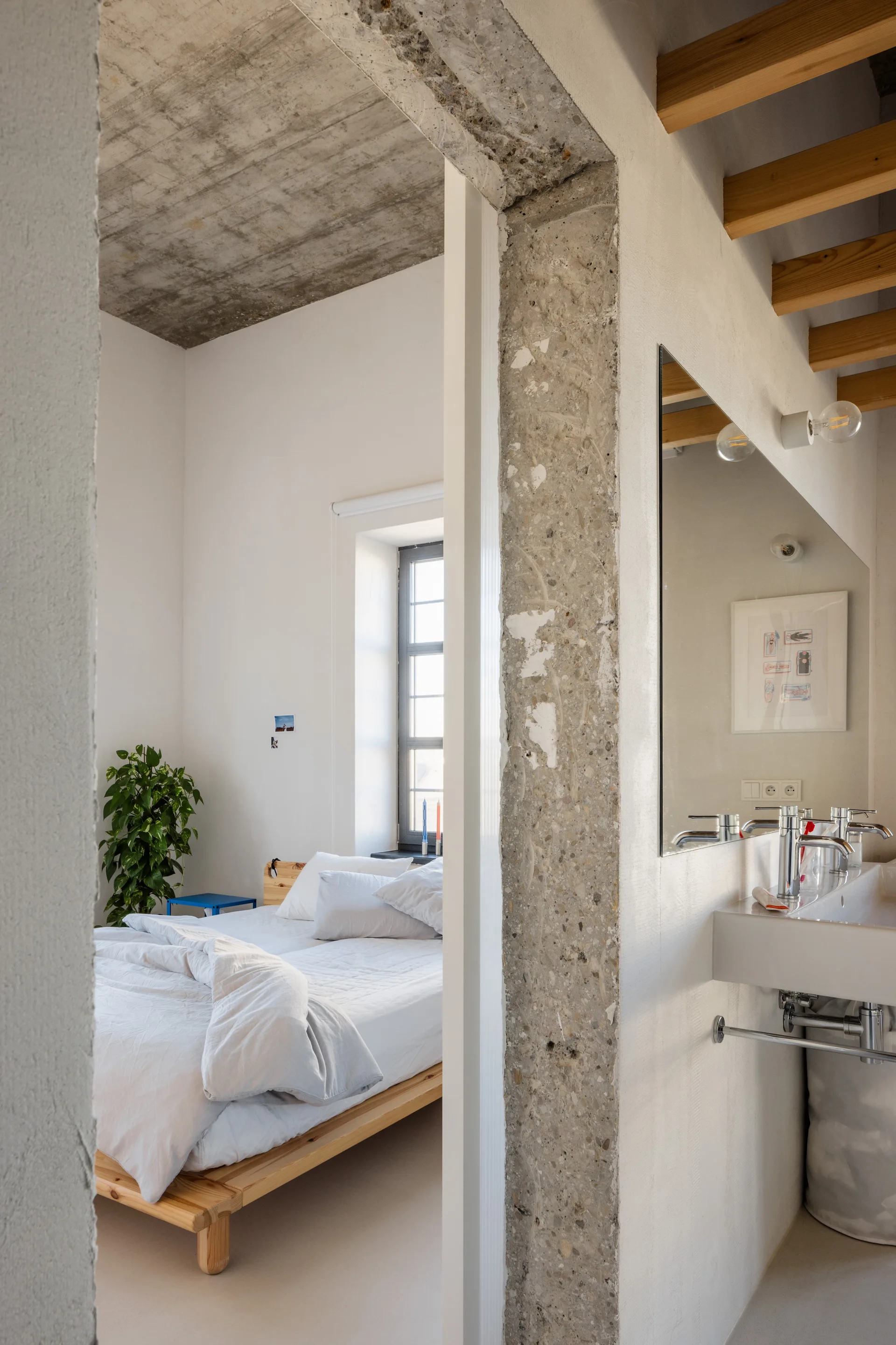 A view from a bathroom with a wall-mounted vanity into a bedroom featuring a wooden platform bed and an exposed concrete ceiling.