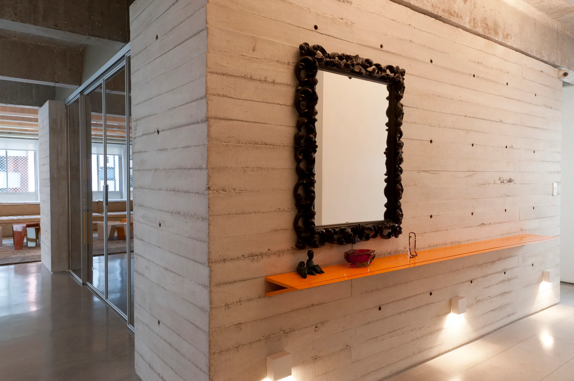 An ornate black-framed mirror hangs above a long, bright orange floating shelf on a textured board-formed concrete hallway wall.
