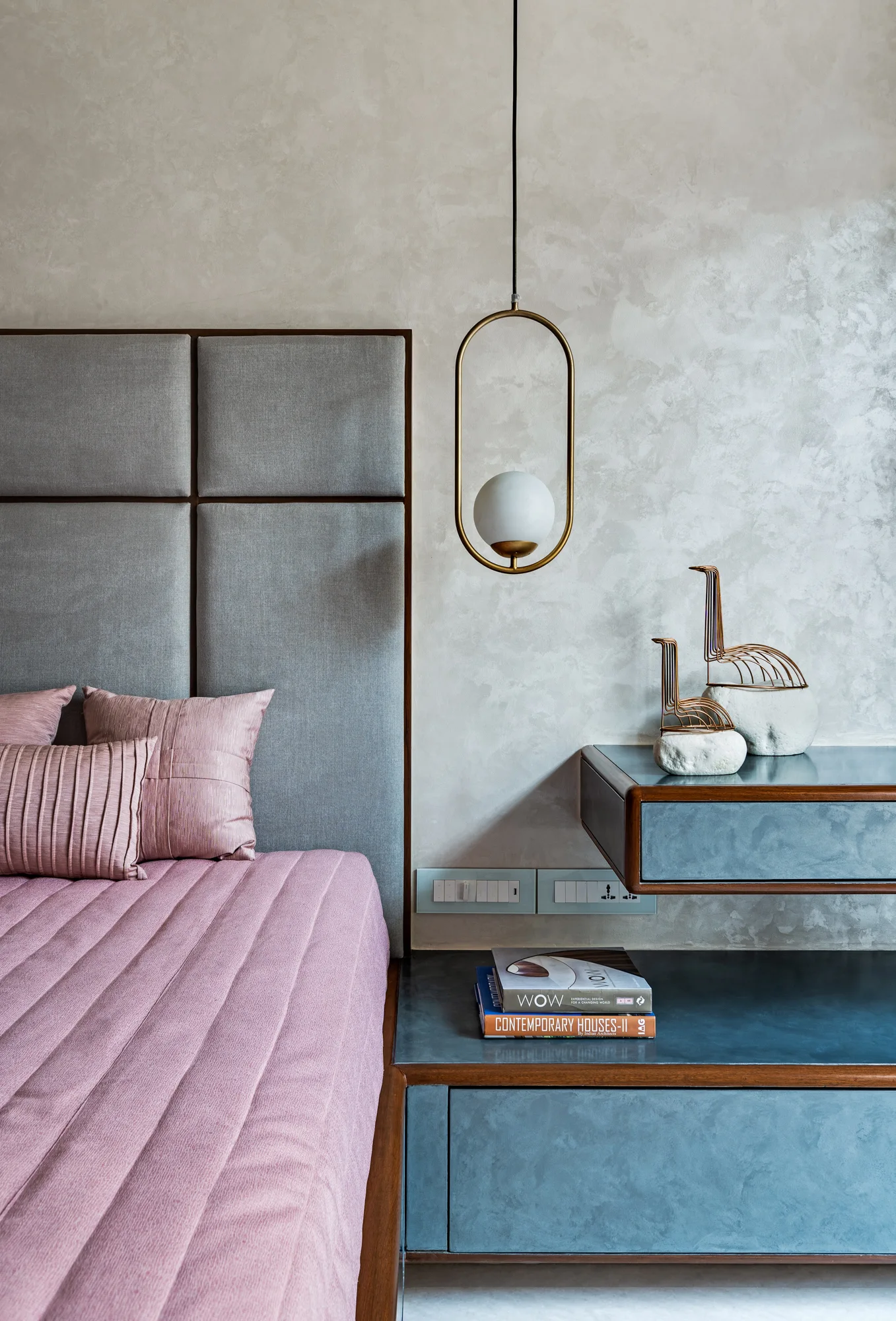 A close-up of a modern bedroom with a pink quilted bed, an upholstered grey panel headboard, and a floating dark grey nightstand.