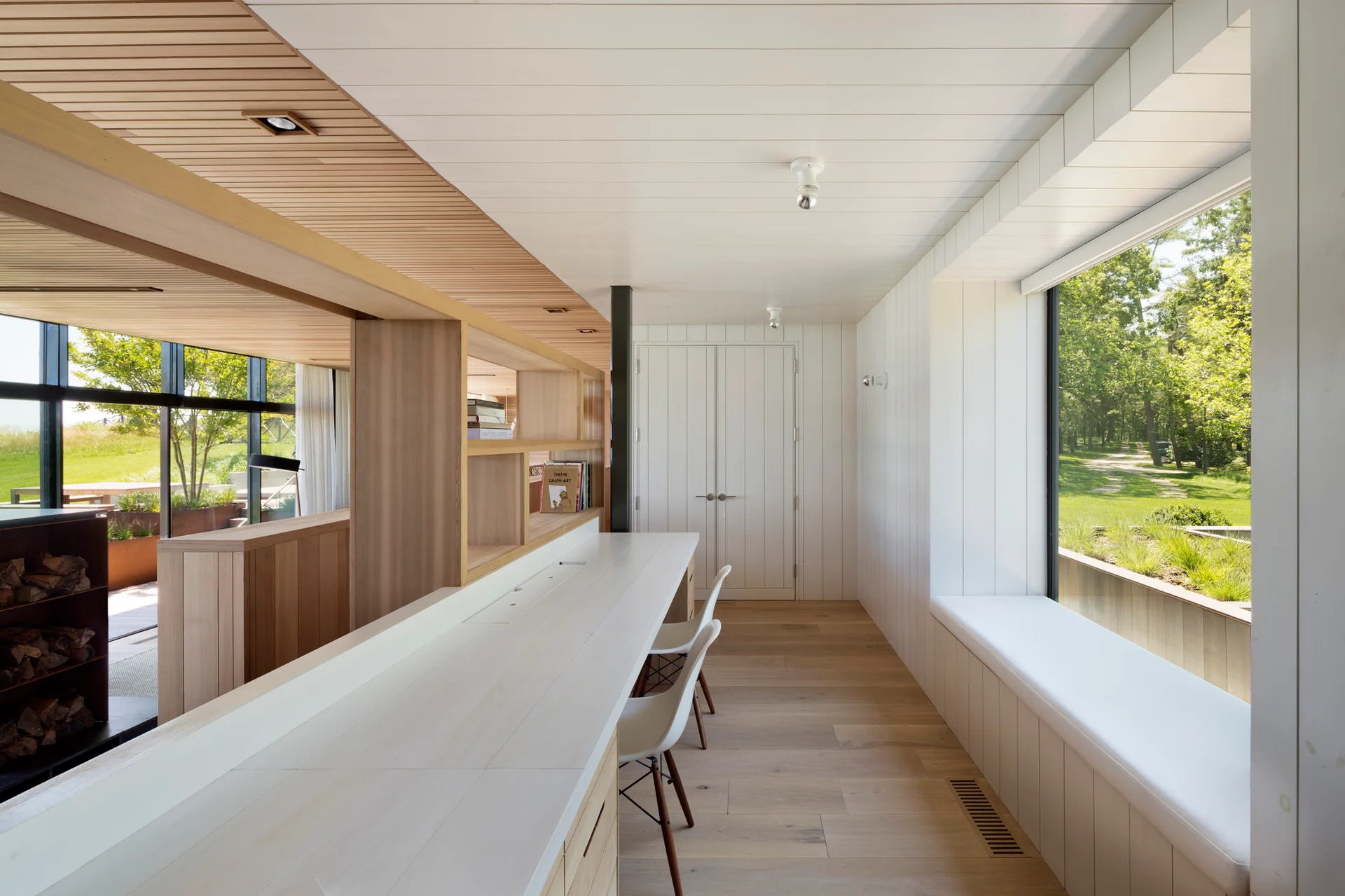 A long white built-in desk with two modern chairs sits opposite a cushioned window bench under a large window overlooking a grassy yard.