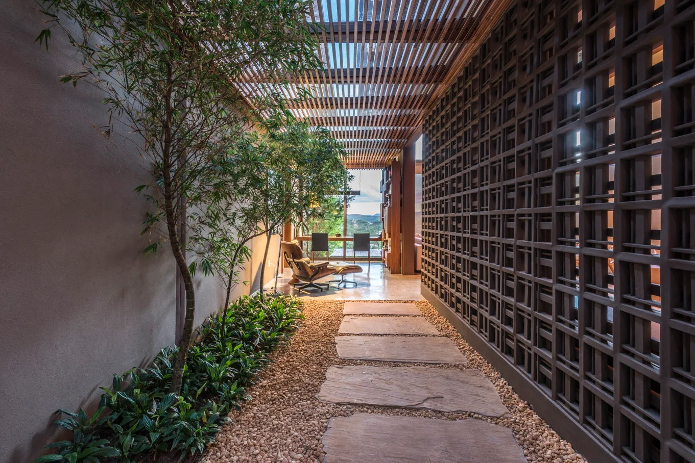 A covered outdoor walkway features a slatted wood ceiling, a decorative geometric block wall, and stone pavers set in gravel leading to a lounge.