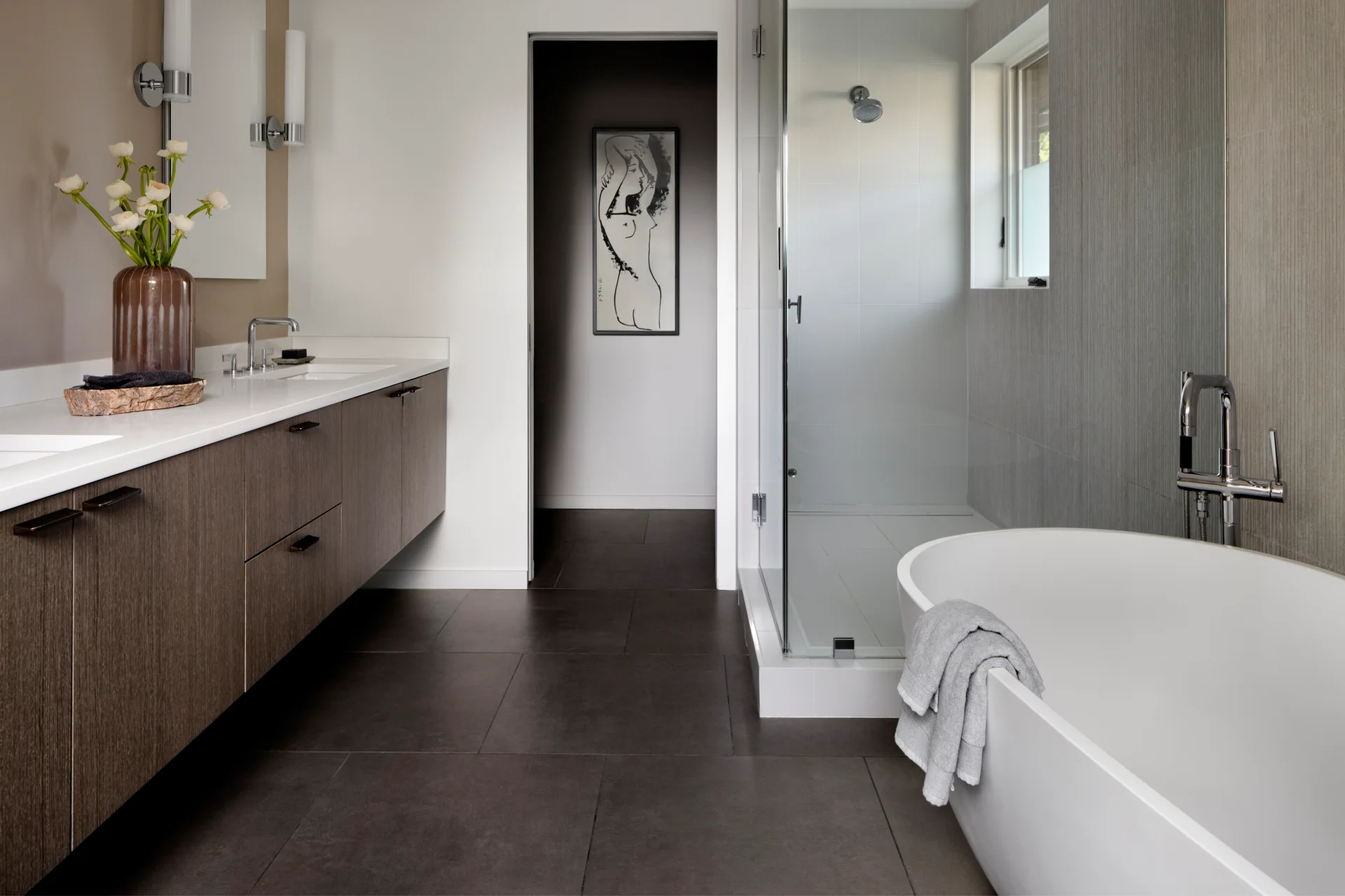 A modern bathroom features a floating dark wood vanity, a glass-enclosed shower, dark floor tiles, and a freestanding white bathtub.