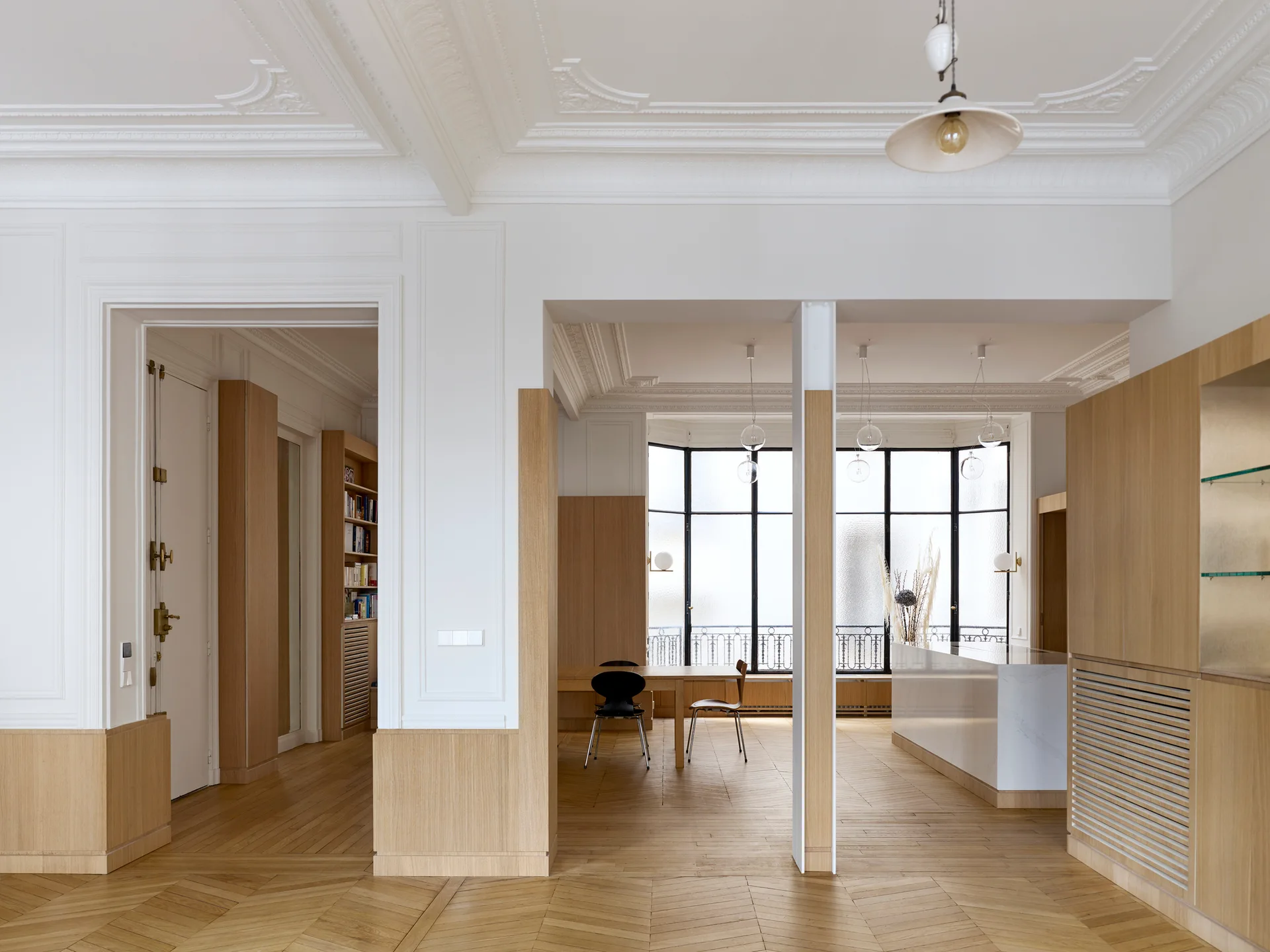 An open apartment dining room featuring herringbone wood floors, white paneled walls with ornate crown molding, and columns wrapped in light oak paneling.