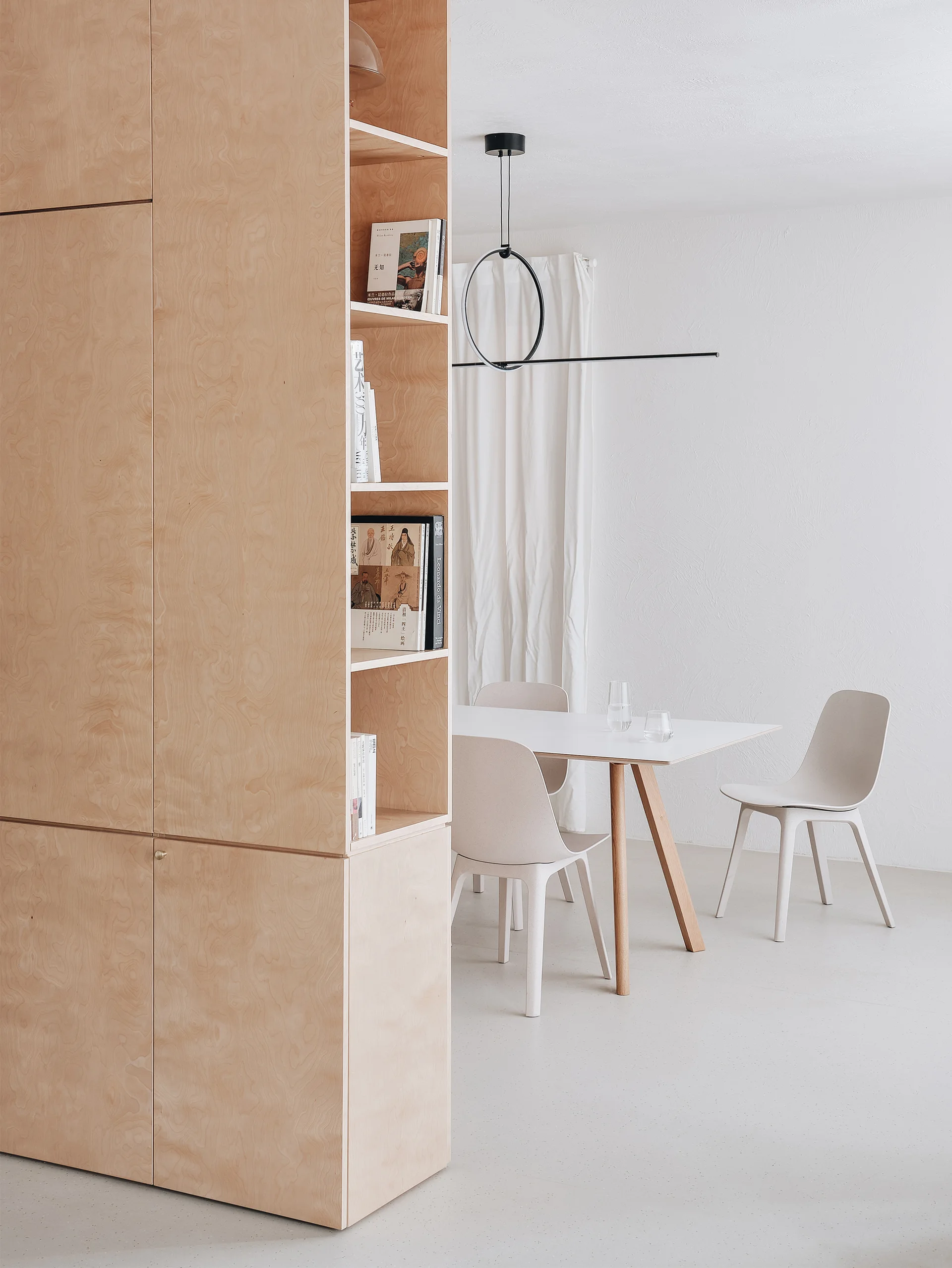 A minimalist dining room featuring a tall plywood shelving unit, a white dining table with molded chairs, and a black circular pendant light.