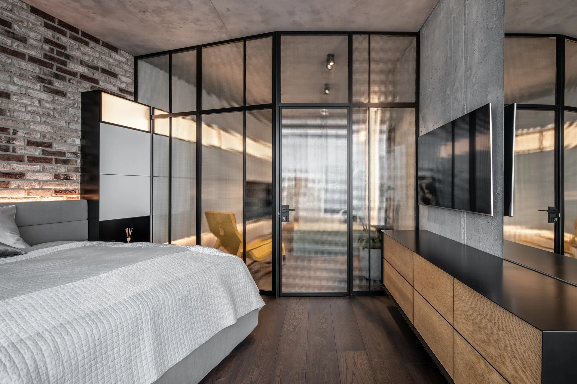 An industrial bedroom features a reeded glass partition with black steel frames, an exposed brick wall, and a low wood dresser under a mounted TV.
