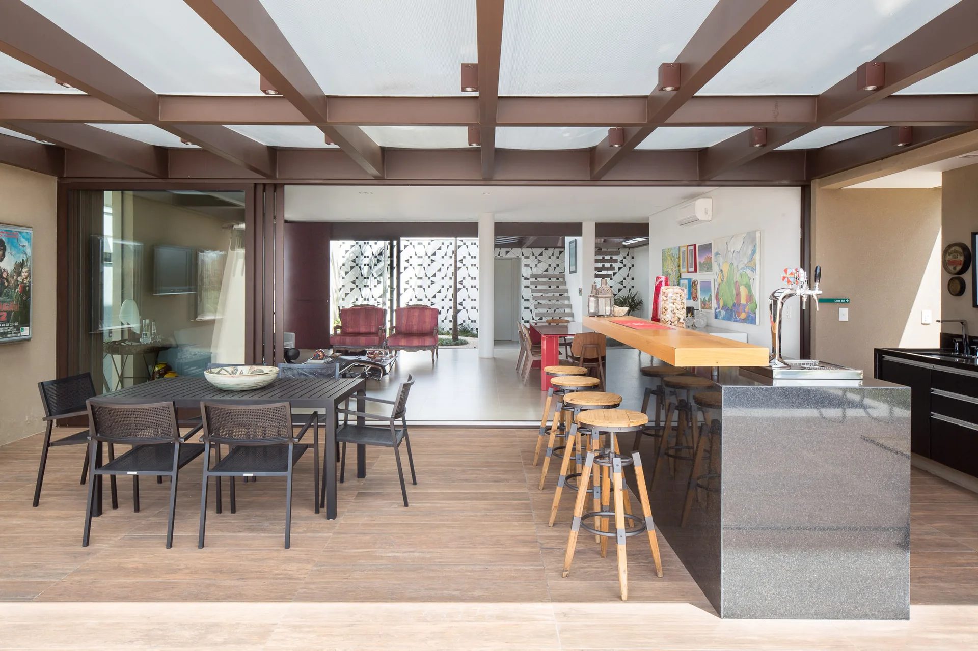 A covered patio featuring a black dining set, a granite bar with wooden stools, and dark ceiling beams that open to an indoor living space.