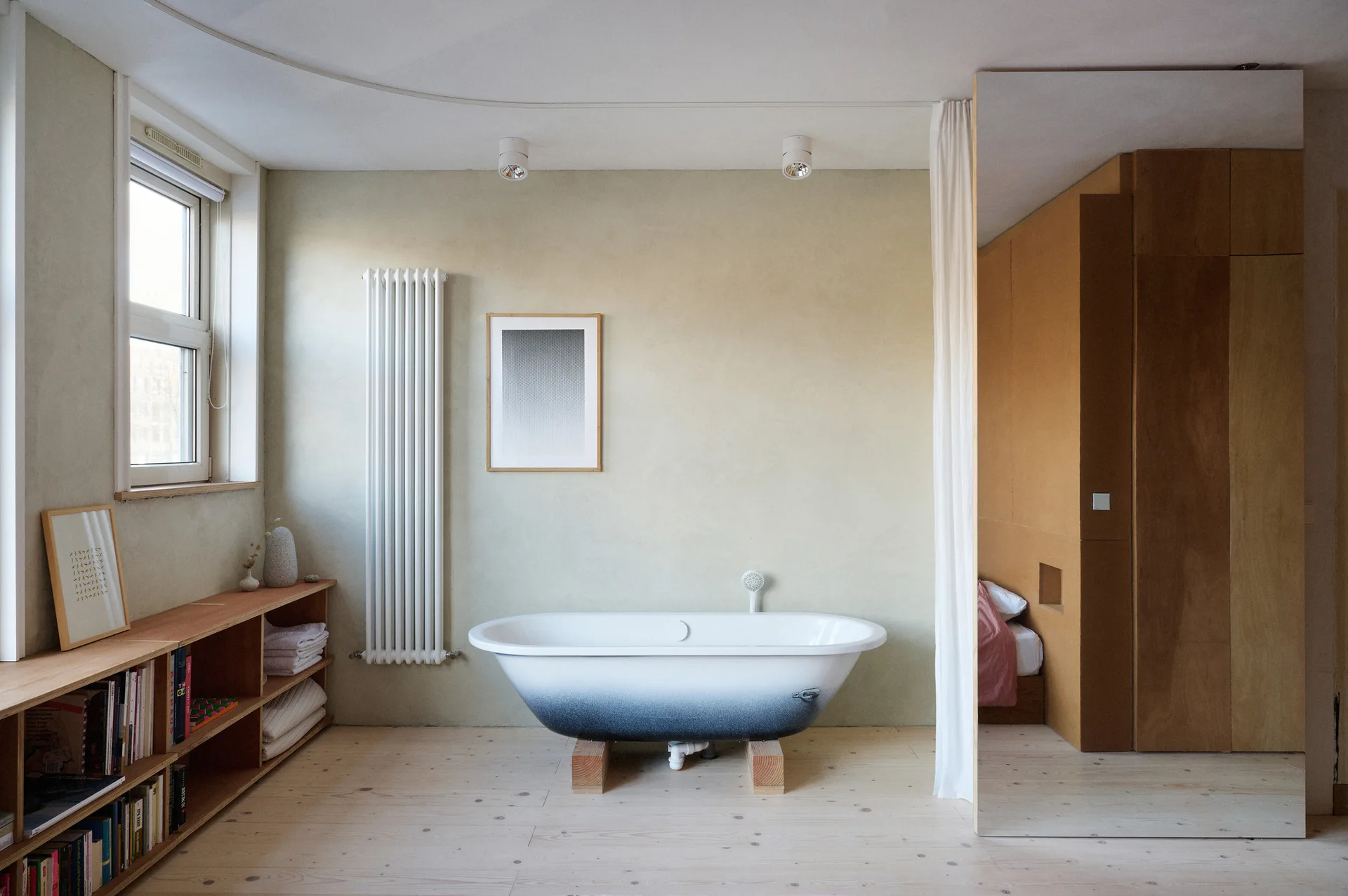 A minimalist bathroom features a white and blue gradient bathtub on wooden supports, a low wooden bookshelf, and a pale green wall.