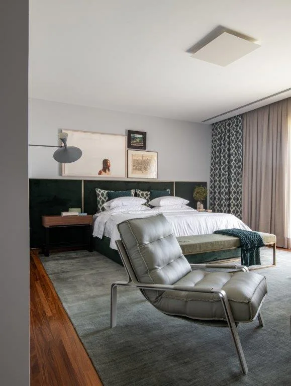 A modern bedroom features a wide forest green upholstered headboard behind a bed with white bedding, a grey leather armchair, and framed wall art.