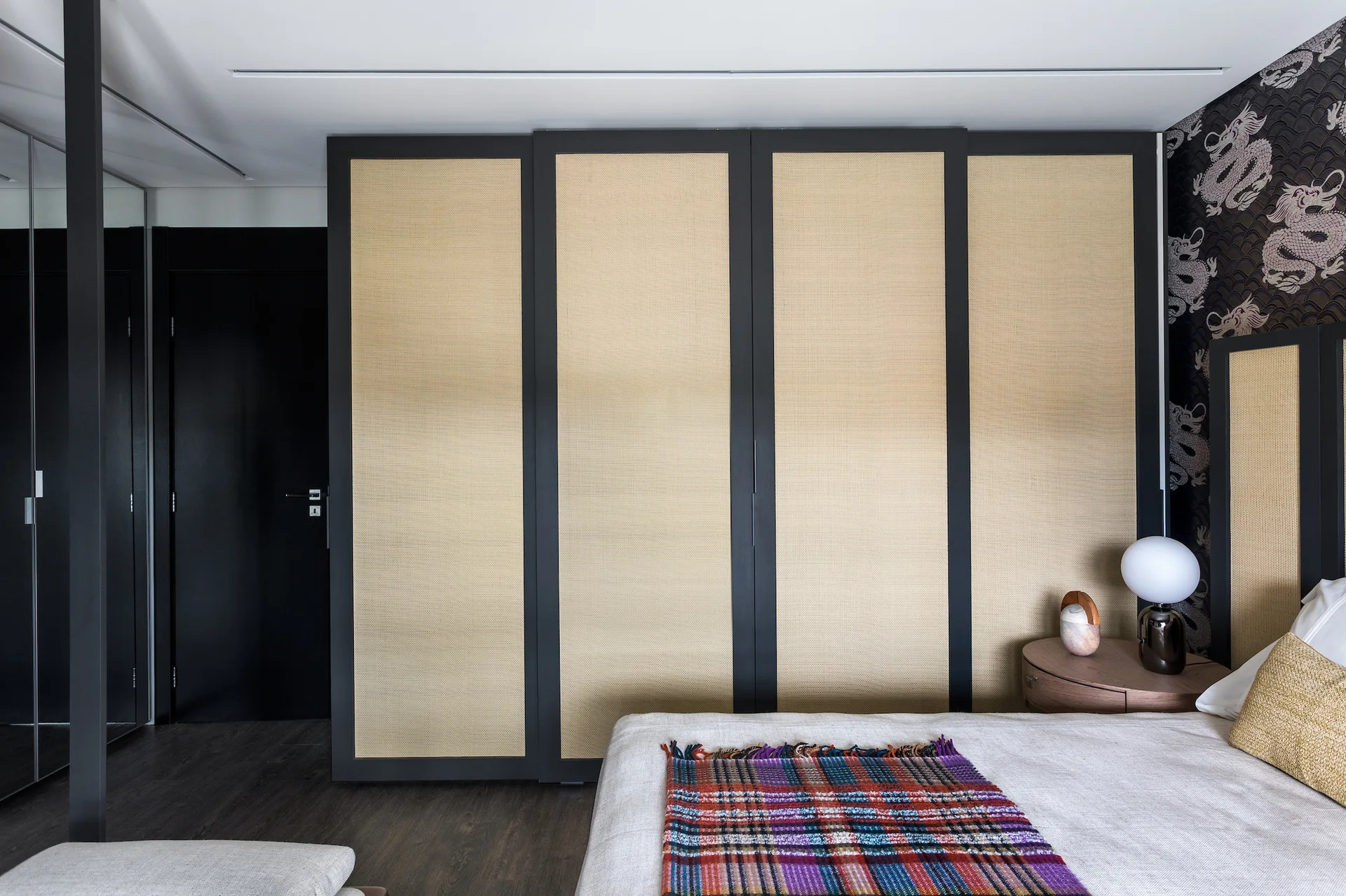 A bedroom features a wardrobe with black-framed cane panels next to a bed with a plaid throw and a dragon-patterned accent wall.