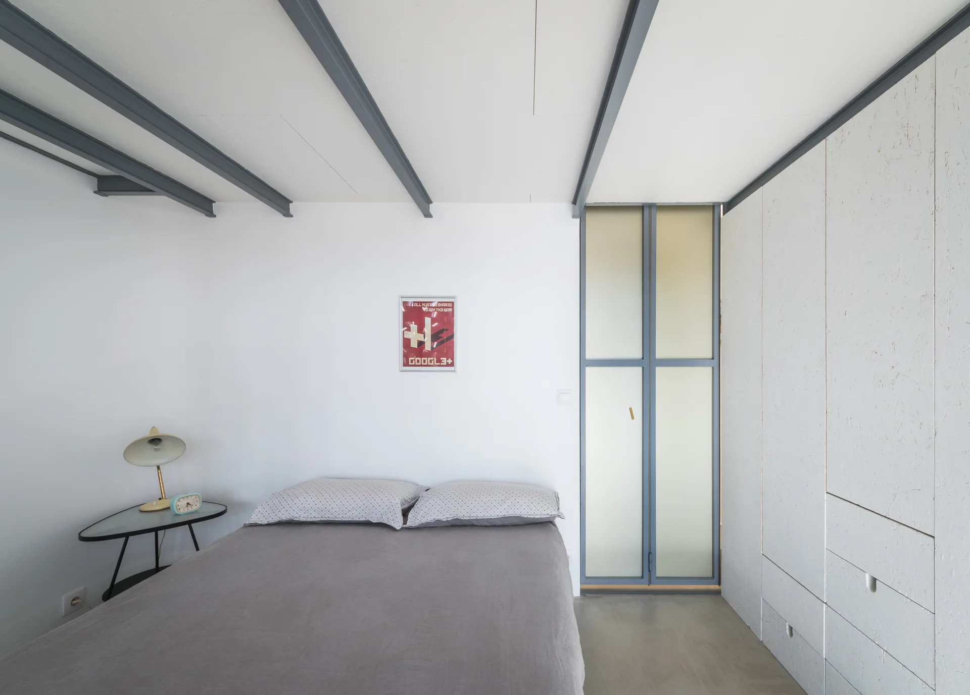 A minimalist bedroom featuring exposed grey ceiling beams, a bed with grey linens, and a white built-in wardrobe.