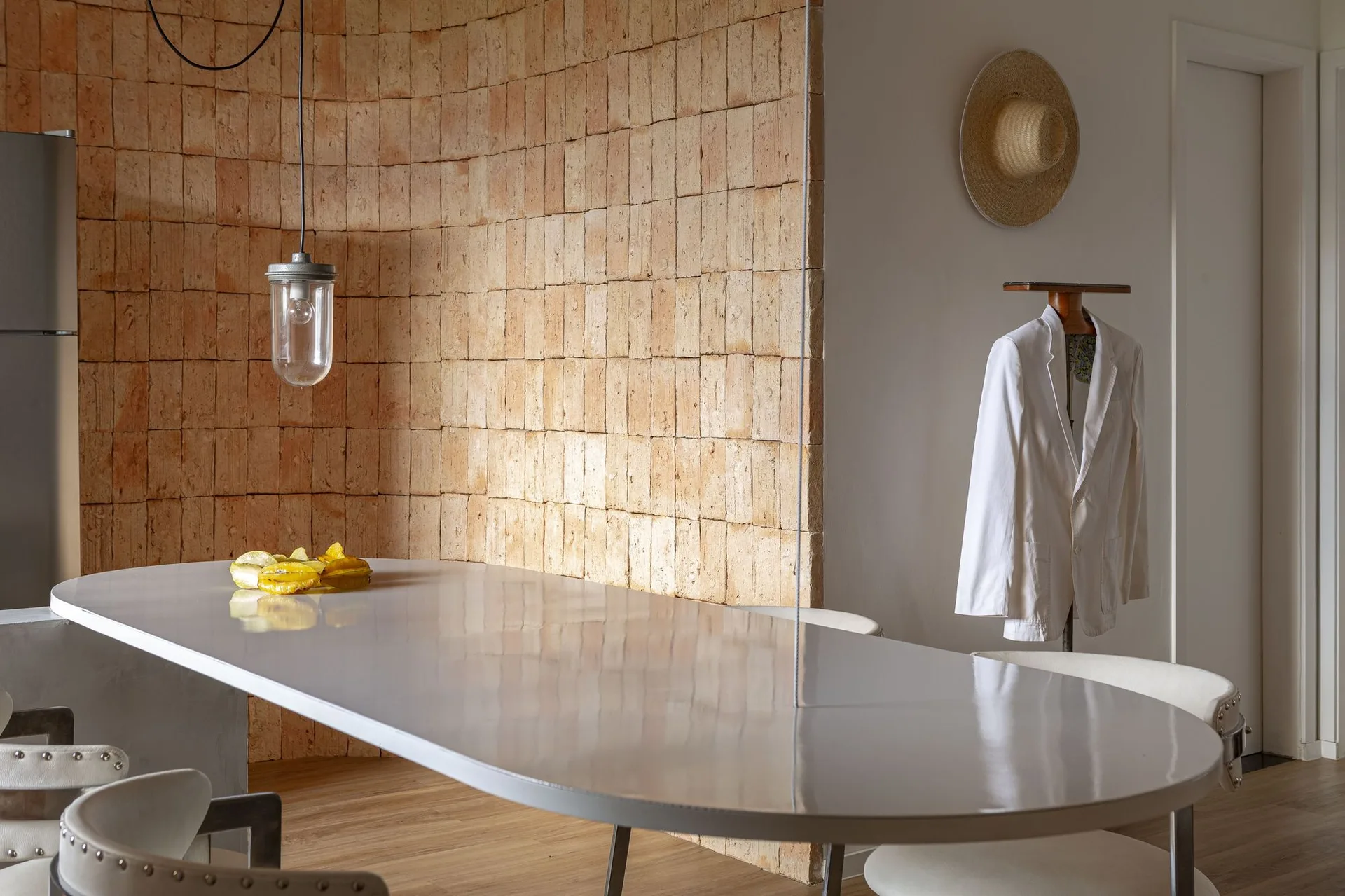 A glossy oval white dining table sits in front of a curved terracotta brick accent wall with a hanging glass pendant light.