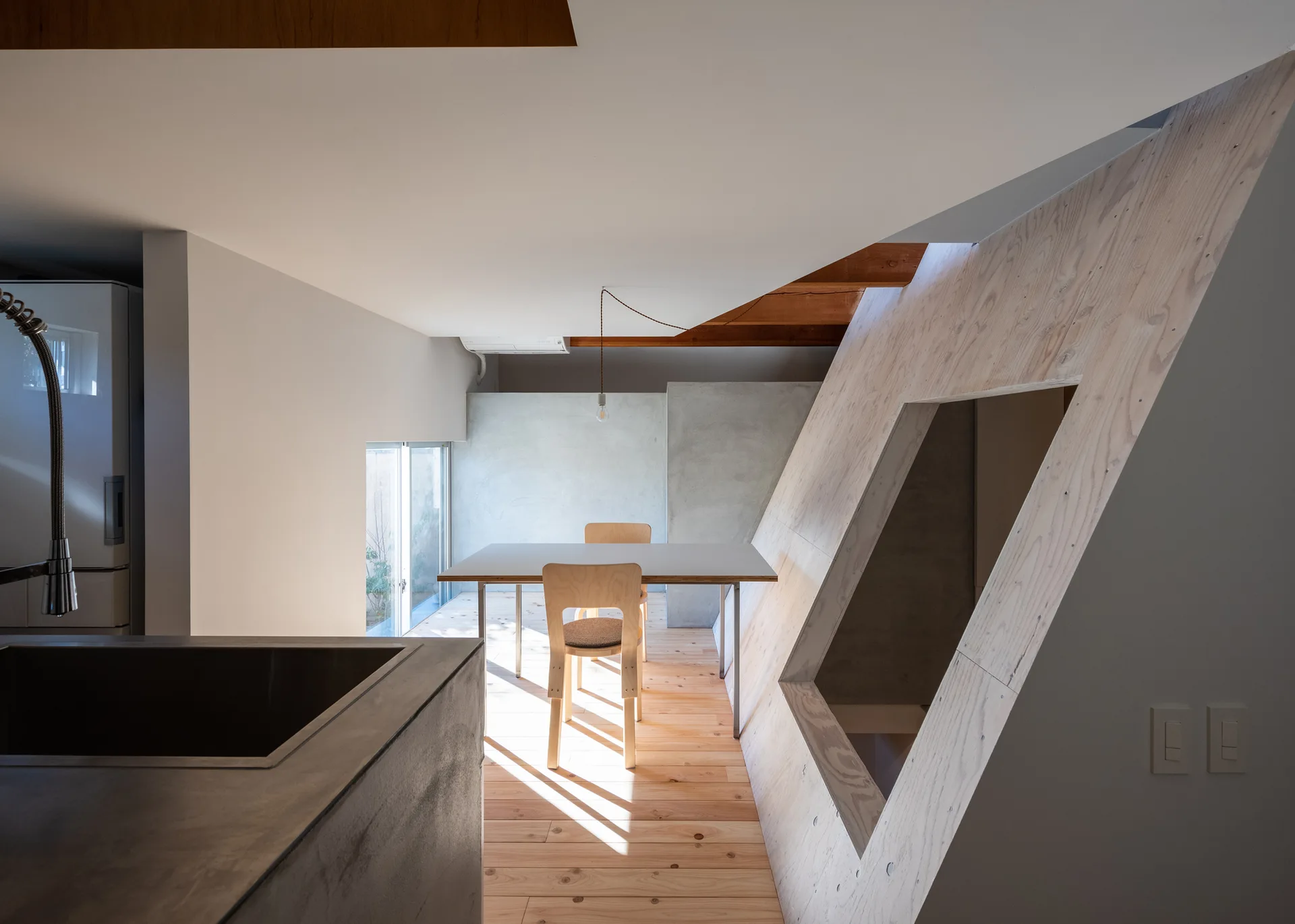A minimalist dining area featuring a light wood table and chairs next to a sloped plywood wall with a geometric cutout.