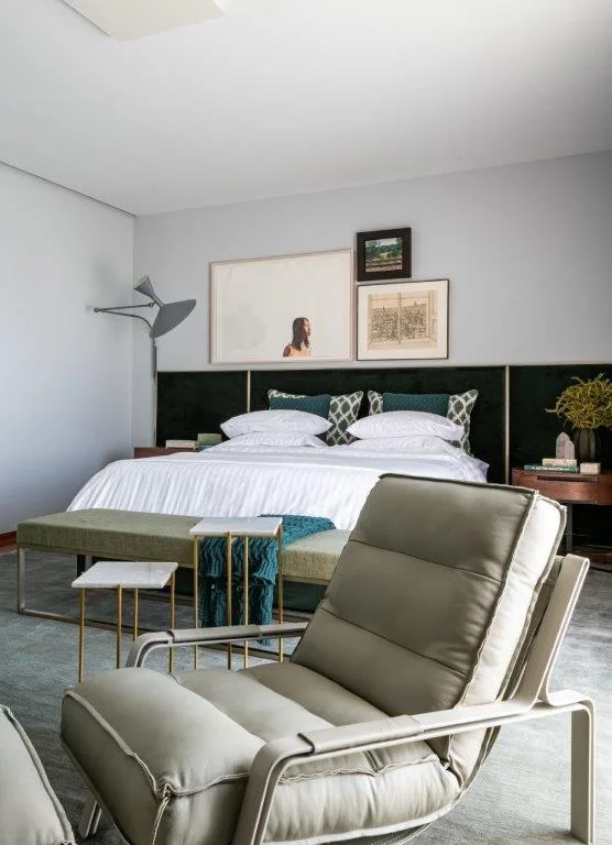 A contemporary bedroom featuring a large bed with a dark green headboard, an olive green lounge chair, and framed artwork on a light grey wall.