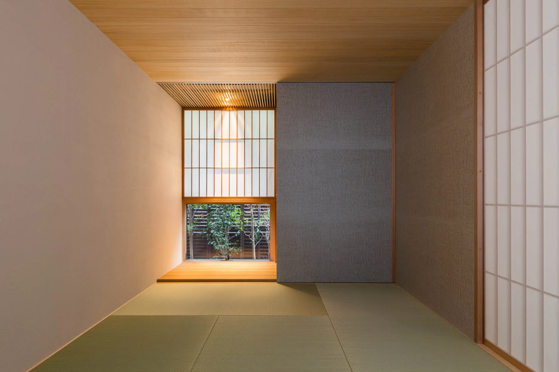 A traditional Japanese room featuring green tatami floor mats, shoji screen walls, a light wood ceiling, and a low recessed window with greenery.