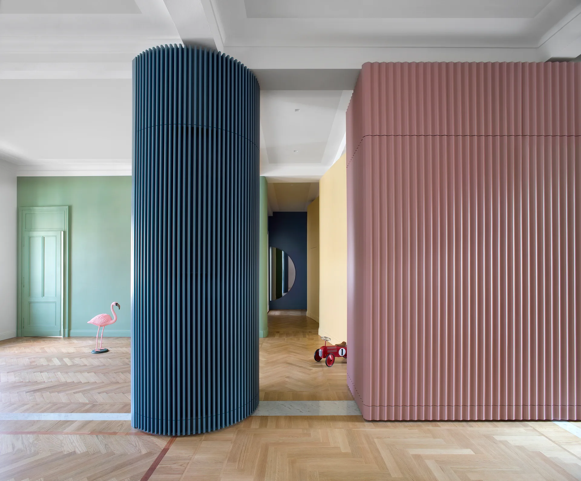 A brightly colored hallway features a blue cylindrical slatted column, a pink slatted partition wall, green walls, and herringbone wood flooring.