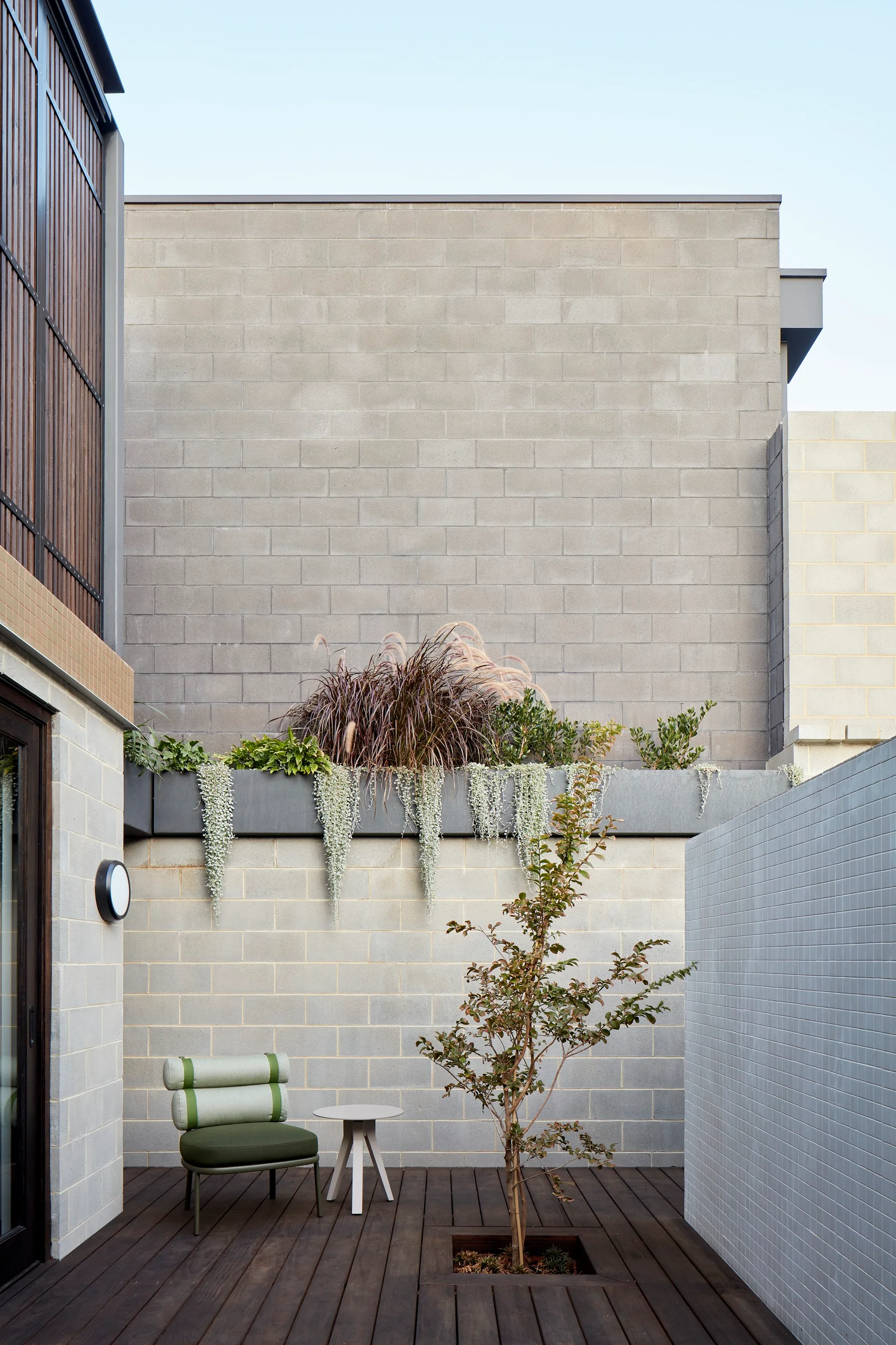 A minimalist concrete block courtyard features a dark wood deck, a modern green armchair, a small side table, and a built-in wall planter.