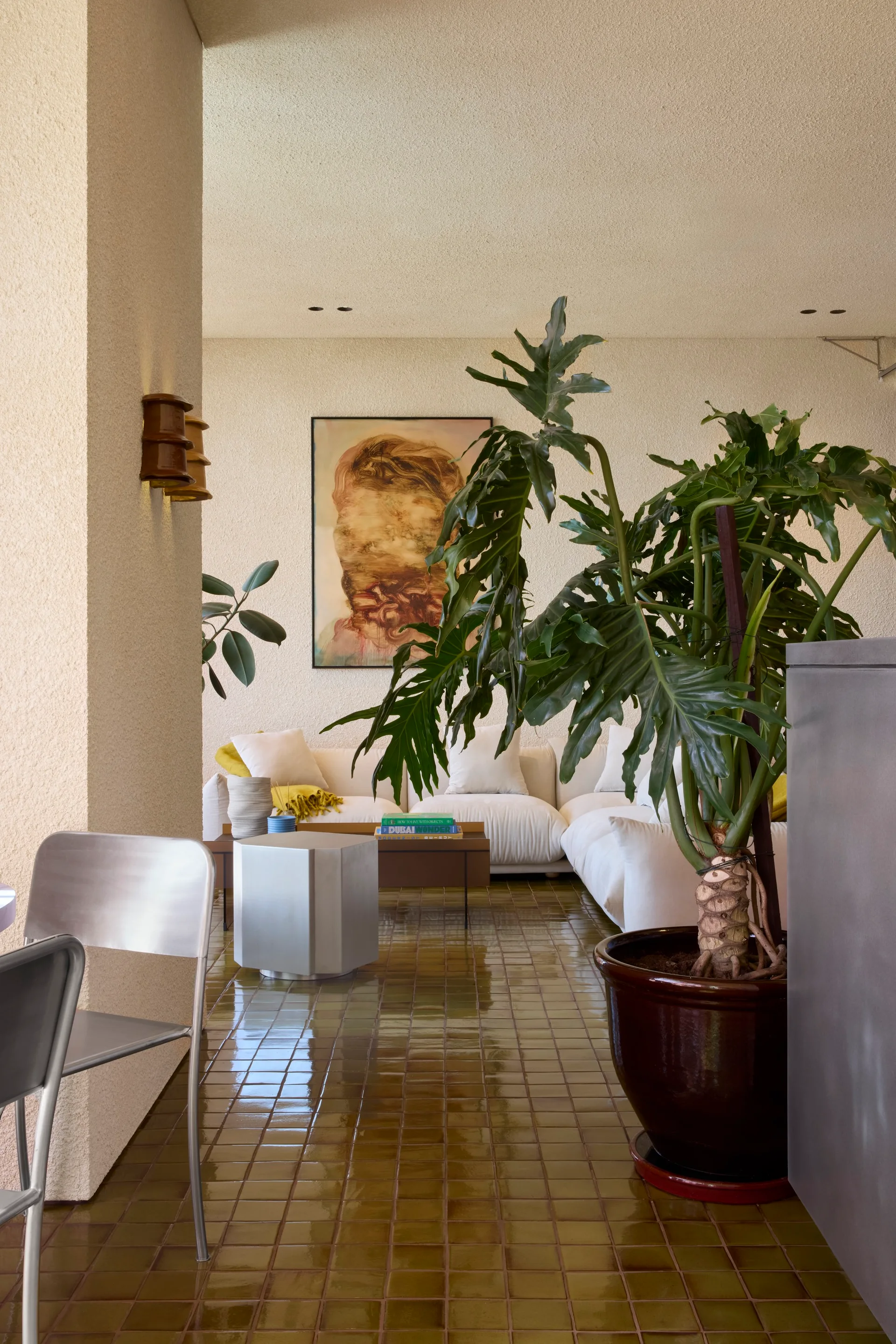 A white low-profile sofa sits on glossy olive green floor tiles next to textured walls, with a large potted plant in the foreground.