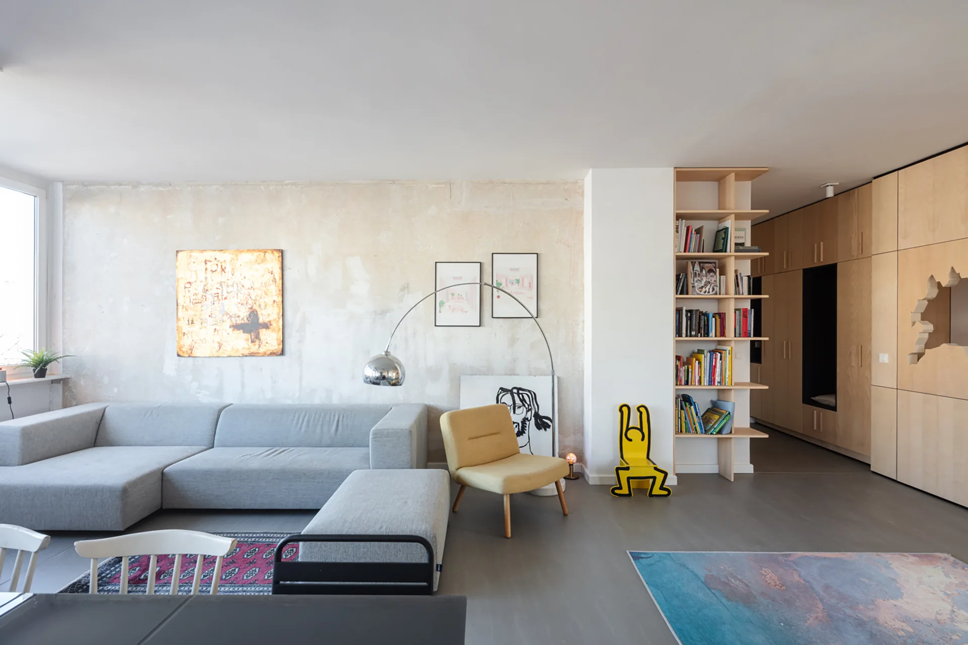 A contemporary living room featuring a grey sectional sofa, a textured plaster wall with art, a yellow accent chair, and light wood built-in shelving.