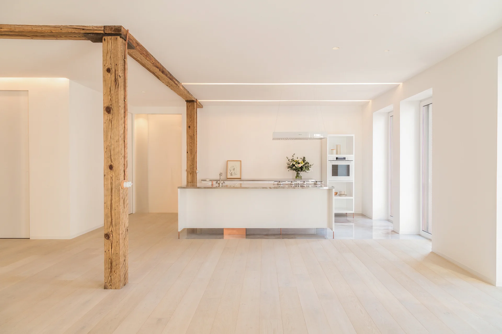 A minimalist white kitchen features a central island, pale wood flooring, and structural reclaimed wood posts and beams.
