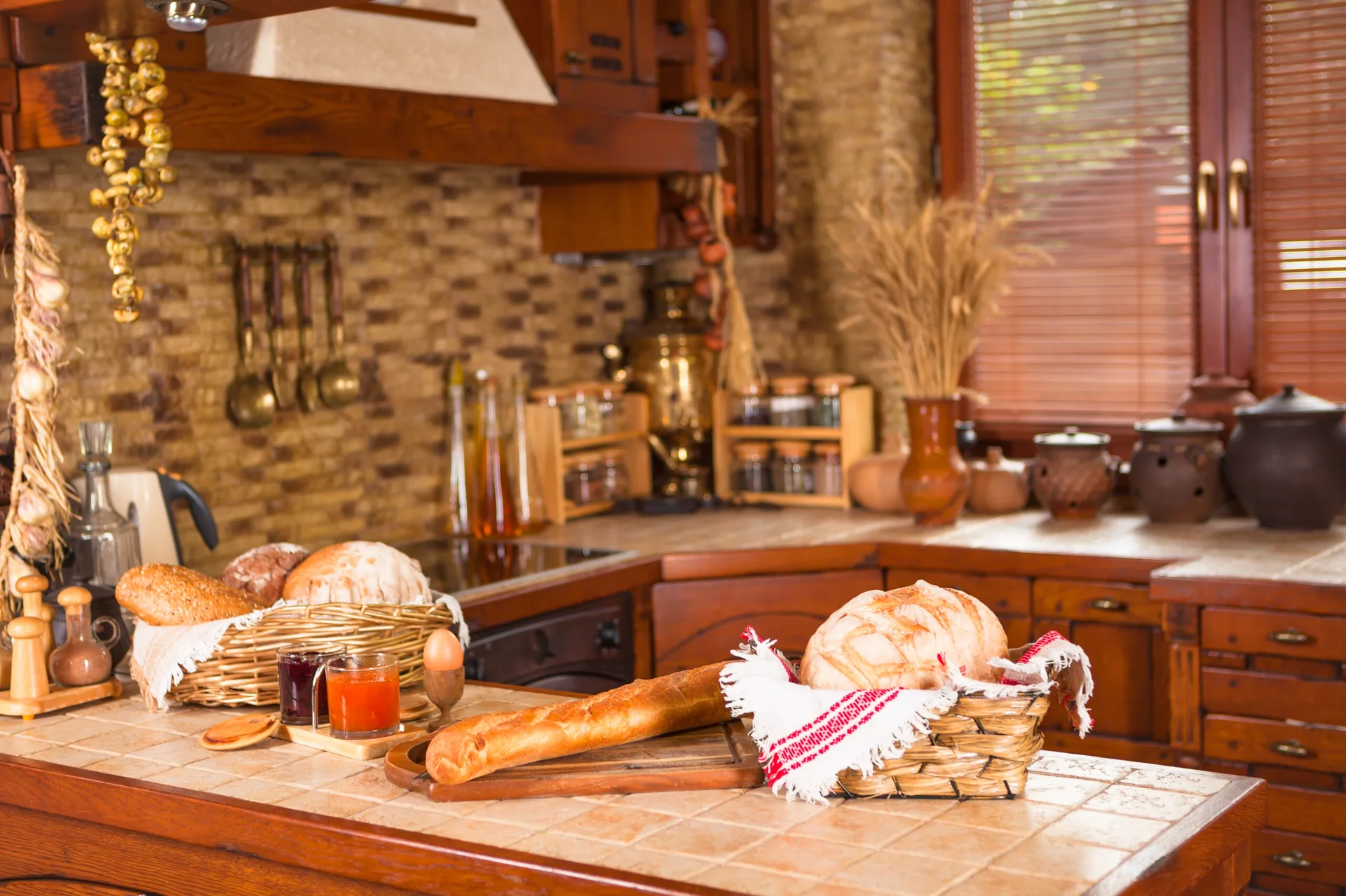 A rustic kitchen features textured stone backsplash walls, dark wood cabinets, and a tile-top island displaying baskets of bread.
