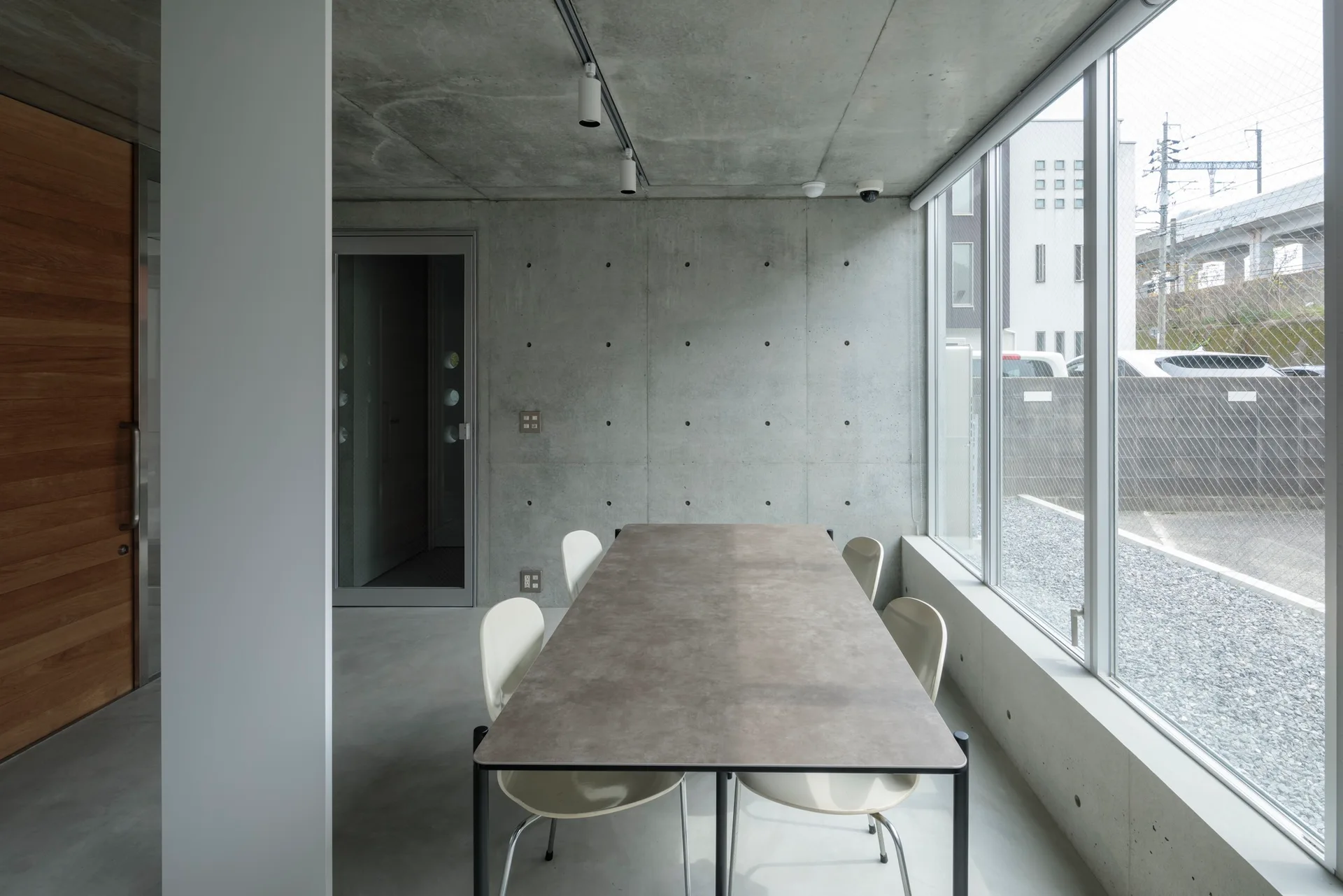 A minimalist dining room features a long rectangular table and off-white chairs next to a large window and exposed concrete walls.