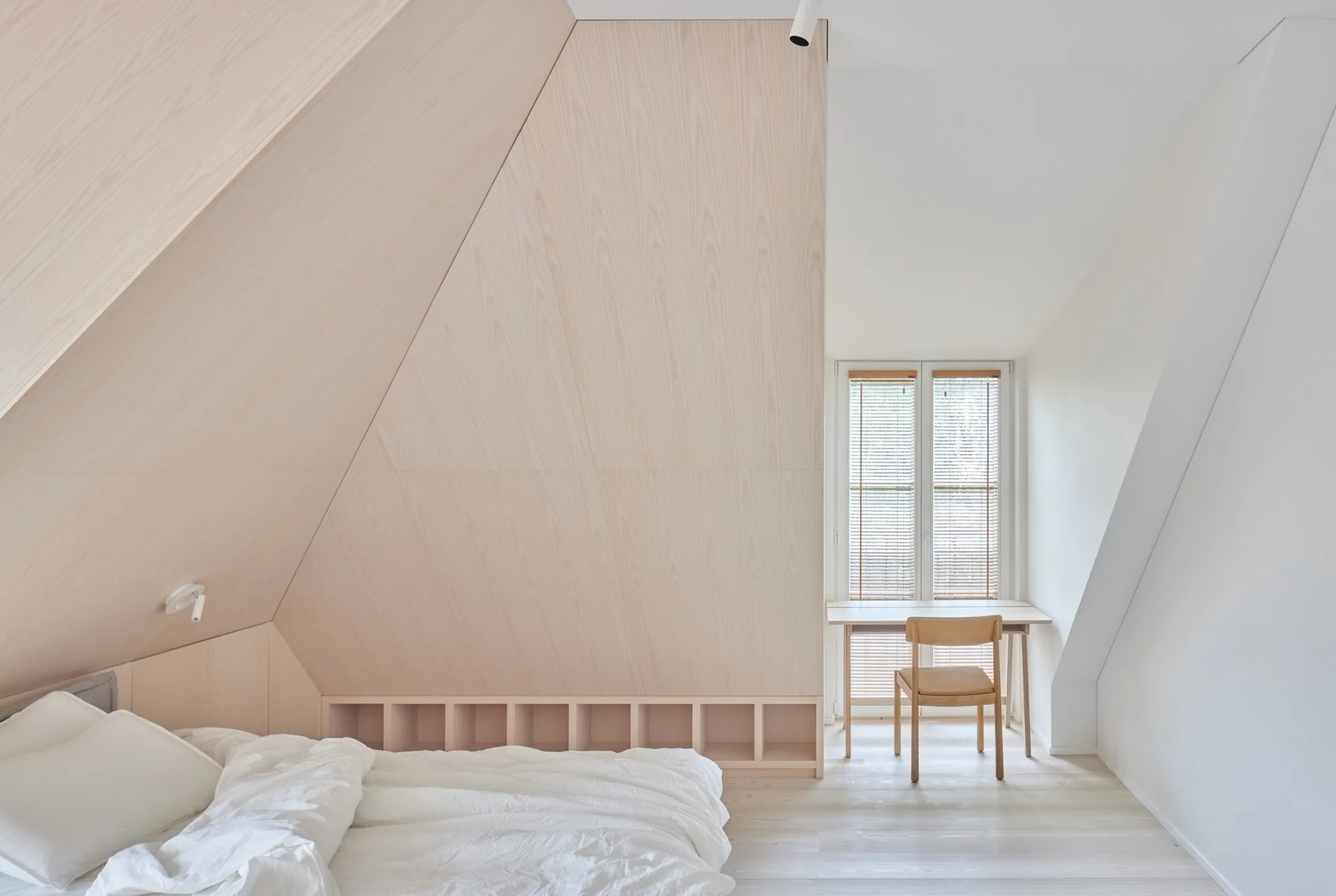 A minimalist attic bedroom featuring a low bed with white bedding, a wooden desk and chair by a window, and sloped wood-paneled walls.