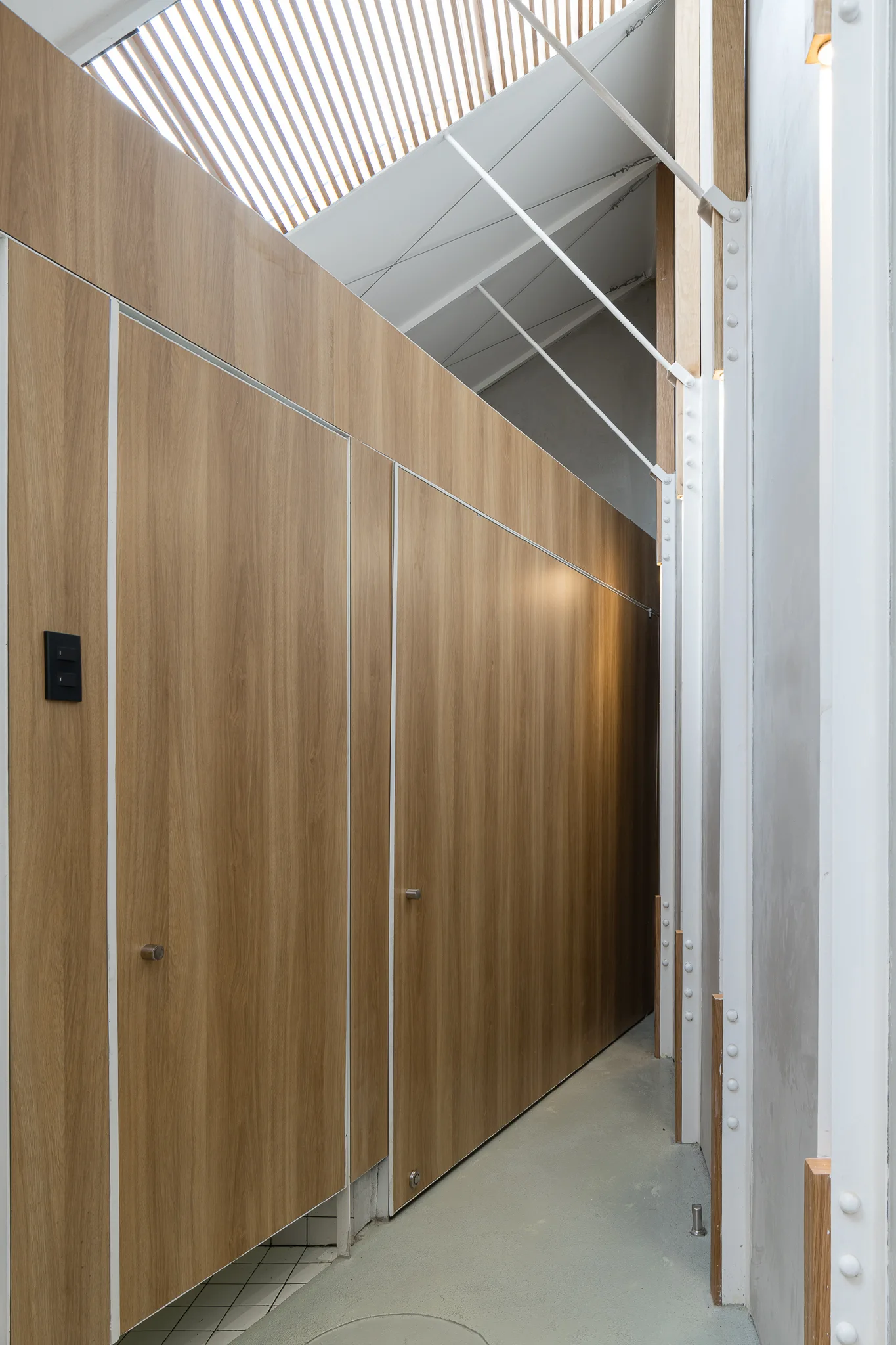 A narrow hallway features smooth wood-toned panel partitions alongside white industrial structural beams with rivets under a slatted overhead skylight.