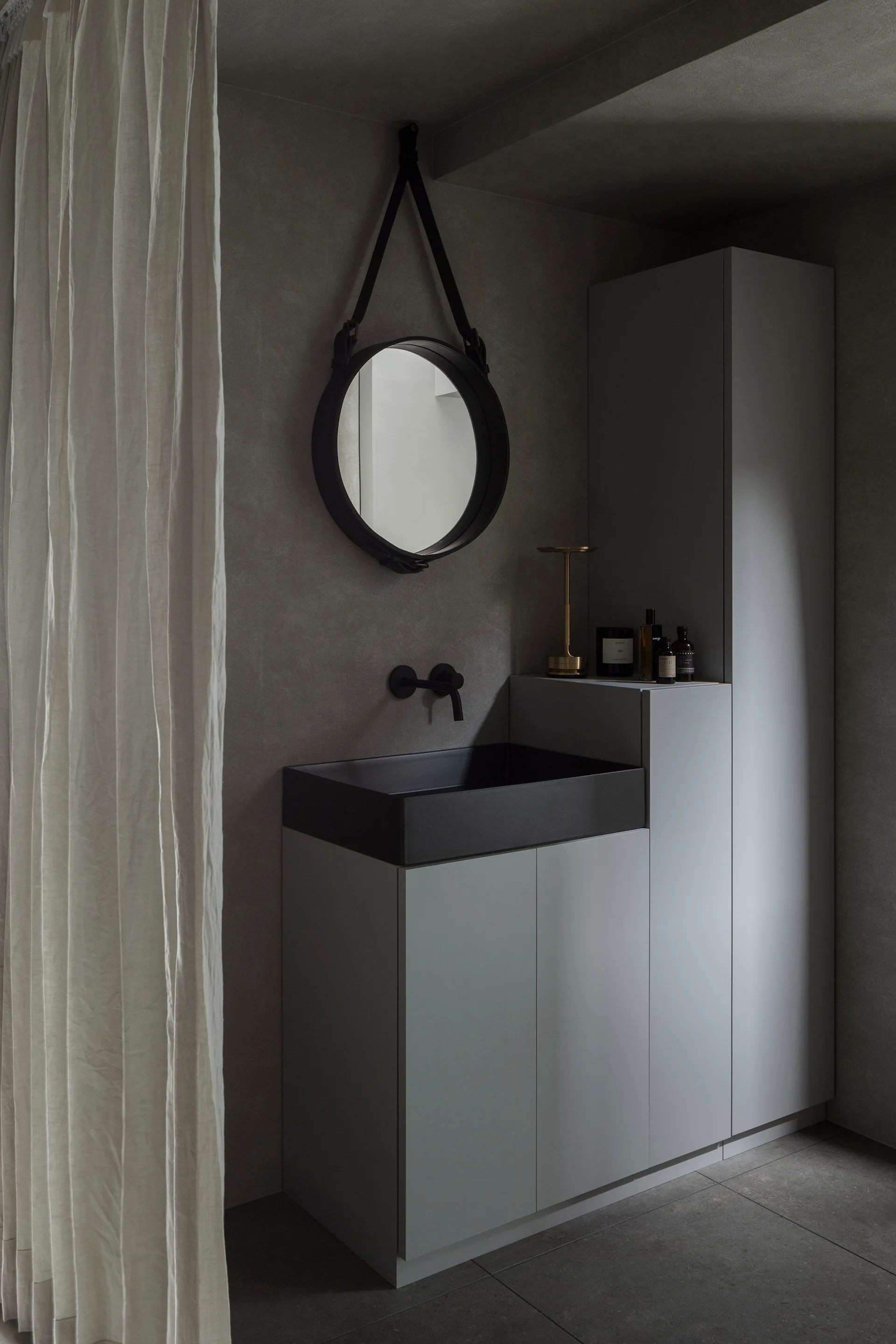 A minimalist grey bathroom features a matte black sink, wall-mounted faucet, circular hanging mirror, and integrated grey storage cabinets.